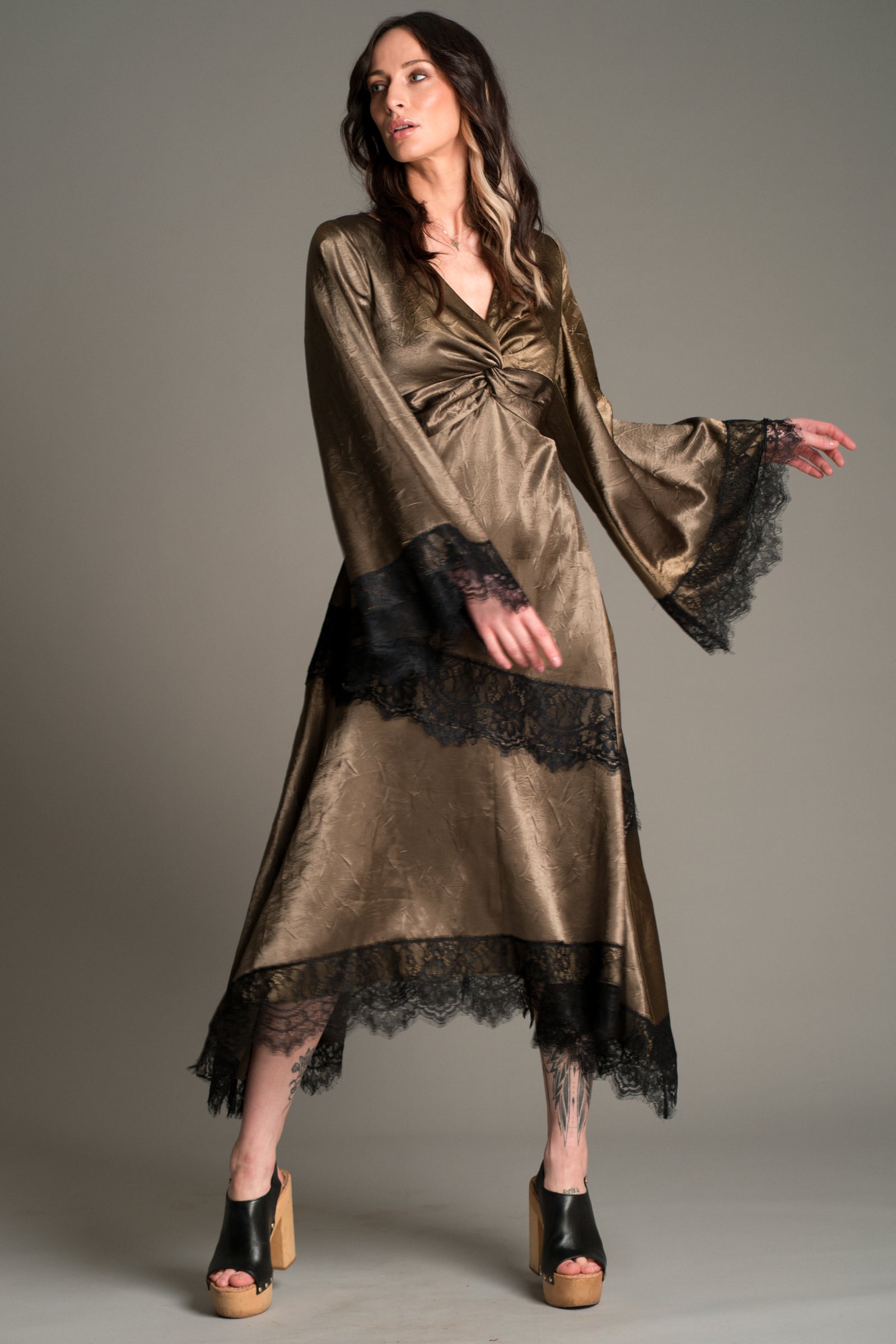 Full‑length view of Victoria in the Little Lies Arabella Satin Maxi Dress in olive satin with black lace trim, featuring flared sleeves and an asymmetric hem, styled with black platform sandals.