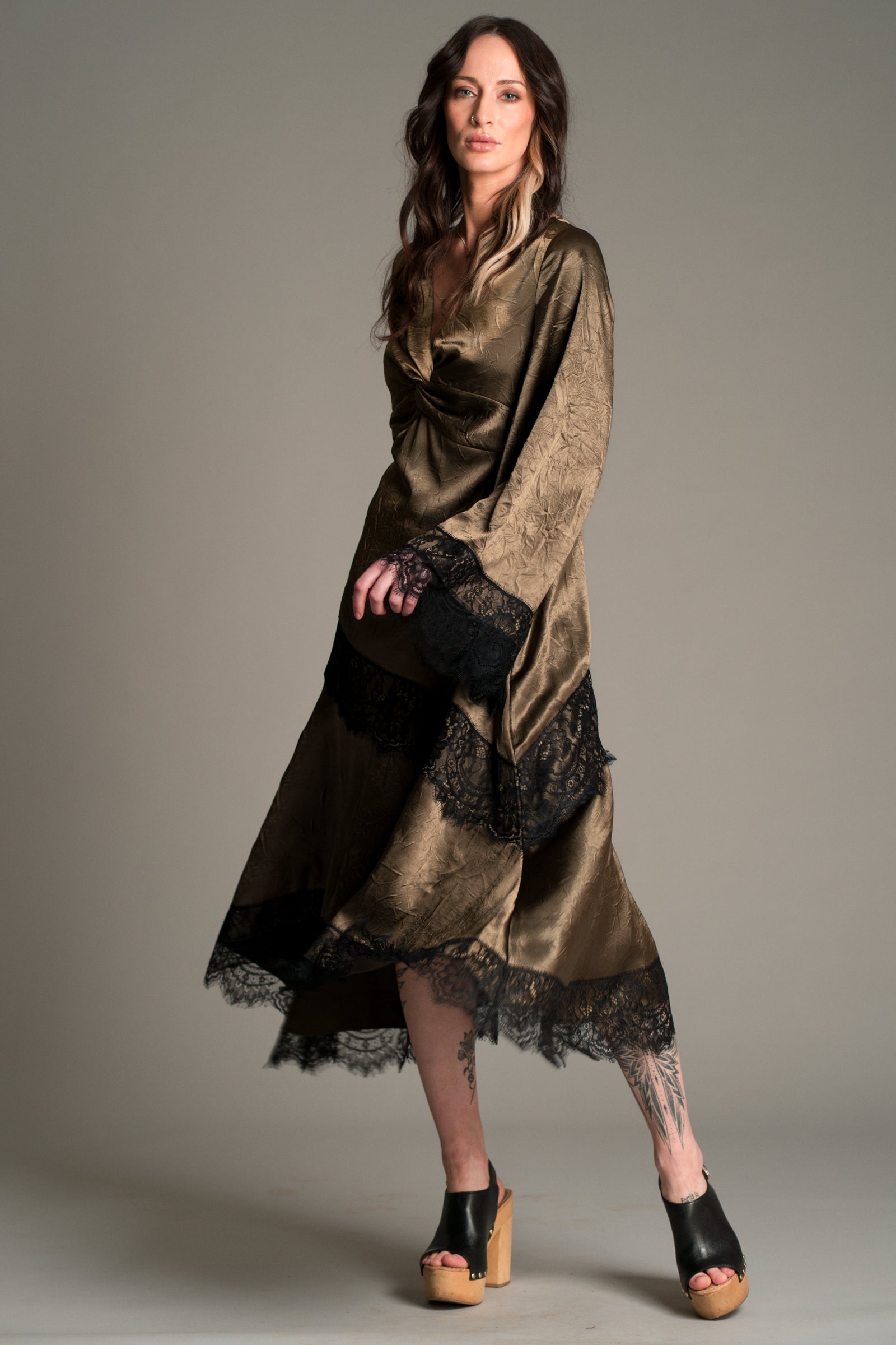 Full‑length view of Victoria in the Little Lies Arabella Satin Maxi Dress in olive satin with black lace trim, featuring flared sleeves and an asymmetric hem, styled with black platform sandals.