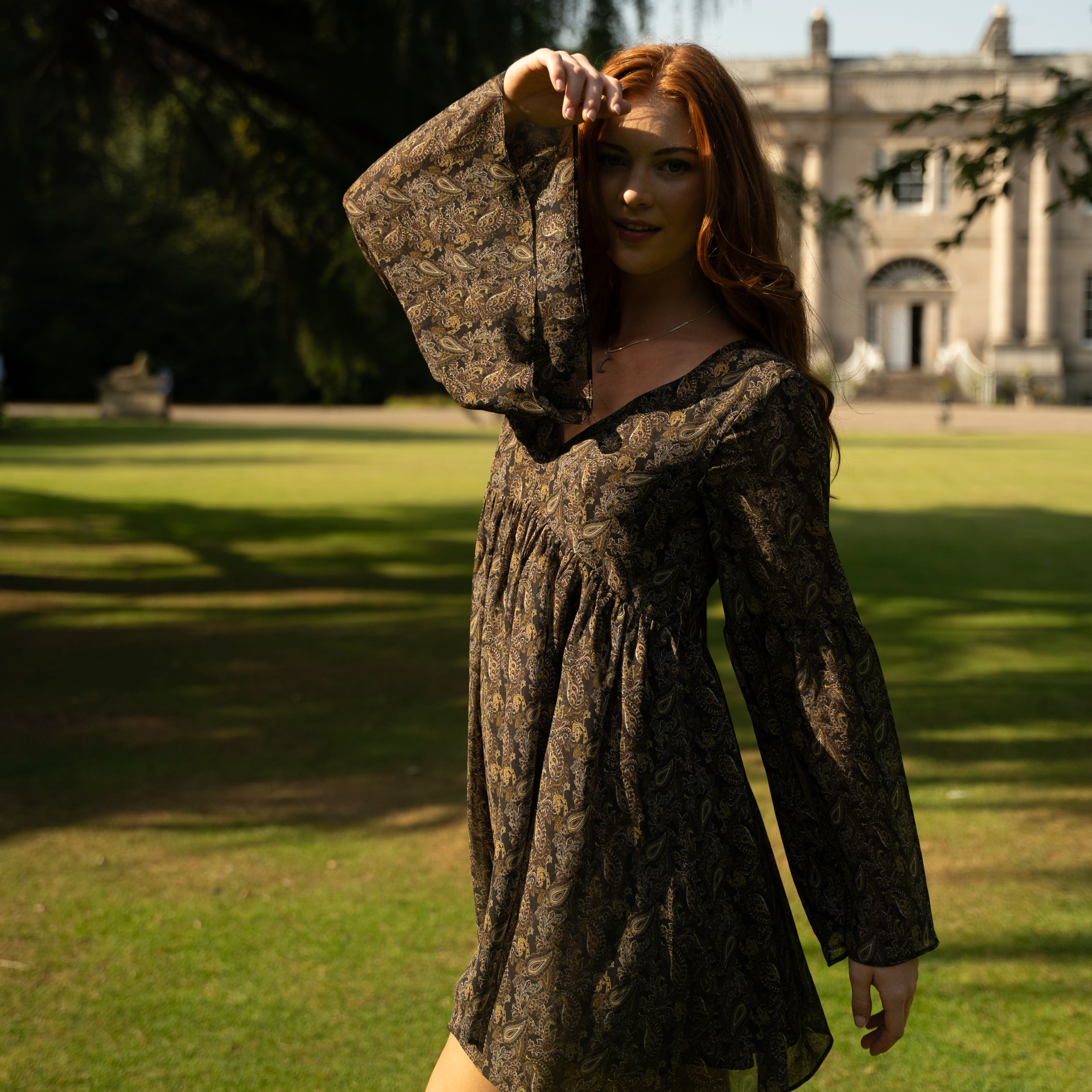 Anna in the Cinnamon Girl Chiffon Paisley Smock Dress standing in estate garden with a building in the background. The Byre at Inchyra