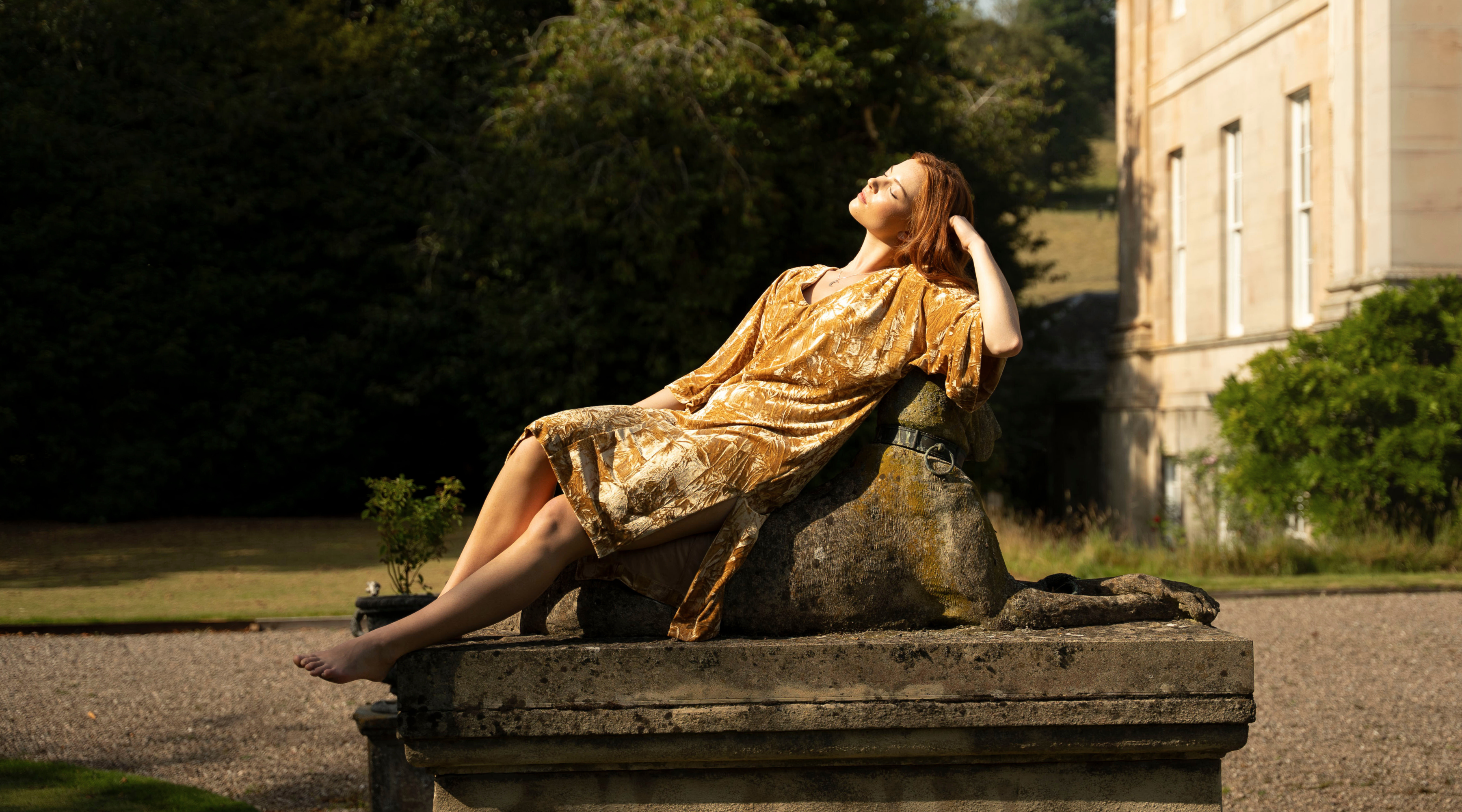 Anna in Rumours Ochre Velvet dress sitting on a stone dog statue at the Byre at Inchyra.