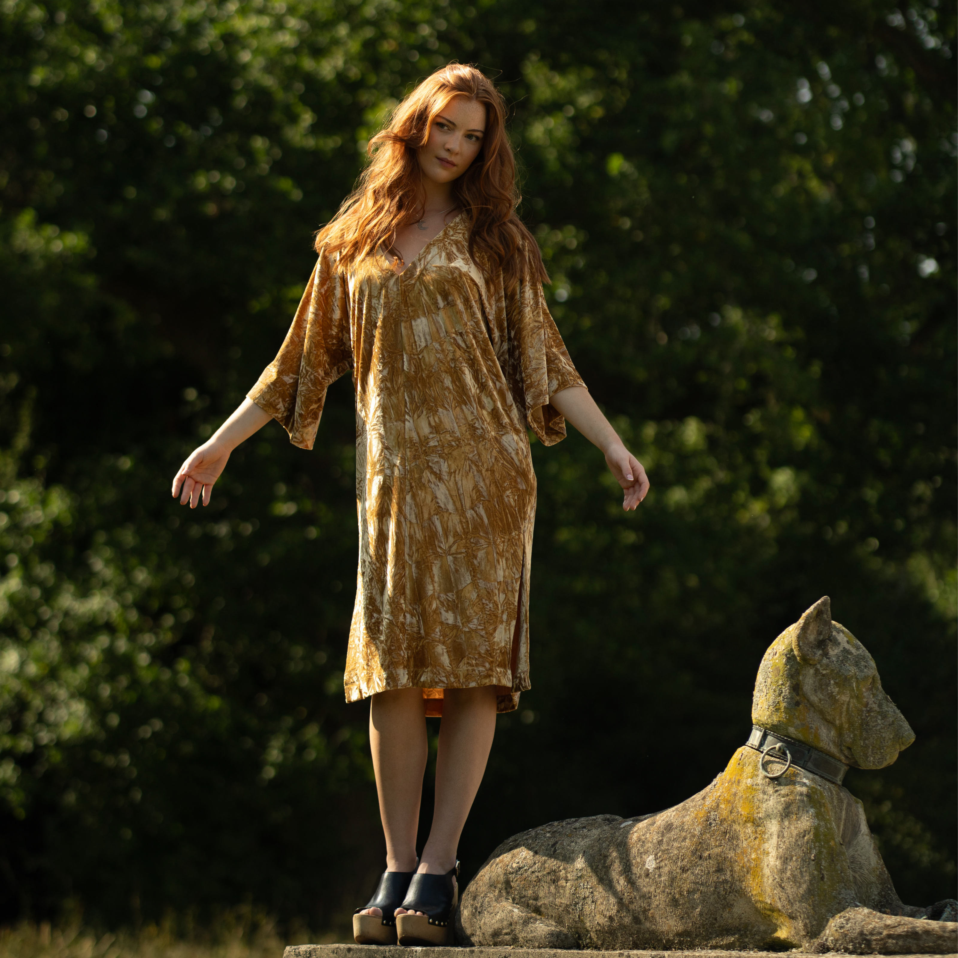 Anna in Rumours Ochre Velvet dress standing on a stone dog statue at the Byre at Inchyra.