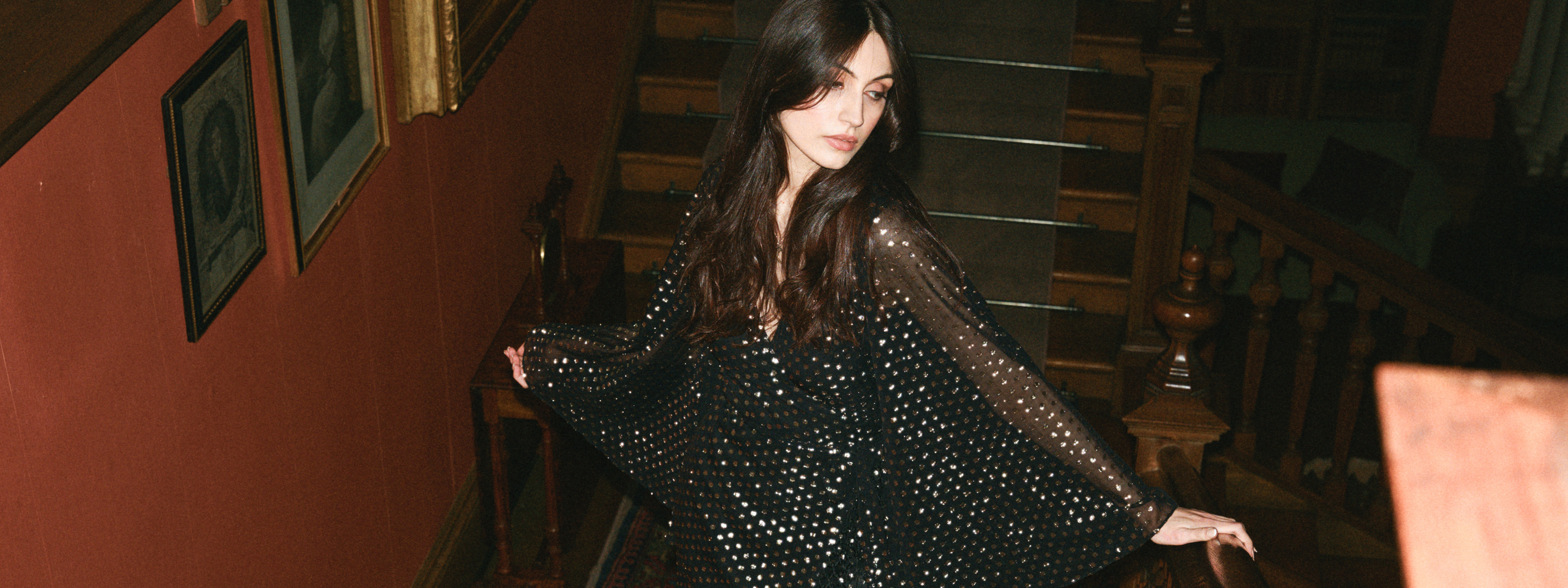 Andie models Little Lies Cosmic Dancer black wrap dress with metallic polka dots and sheer bell sleeves styled on a grand staircase – glamorous evening look