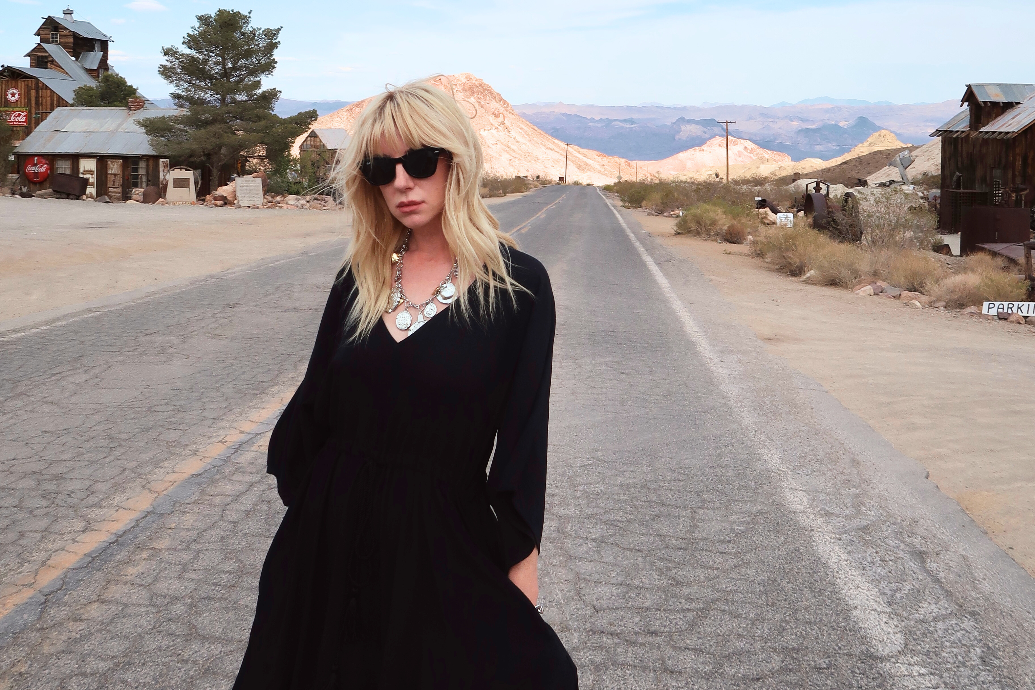 Model in black Blackbird Midi Dress standing on a Western road with rustic buildings and mountain backdrop.