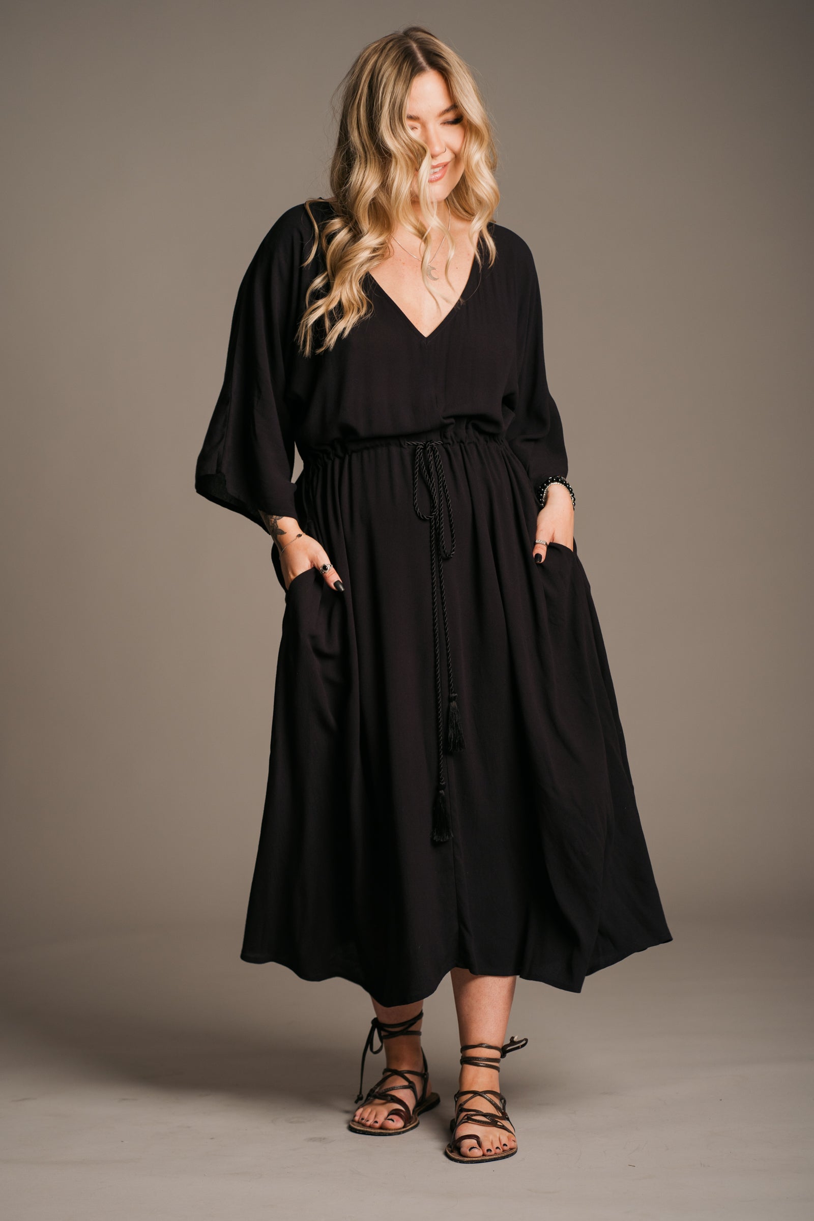 Model Erin wearing the Little Lies Blackbird Tie Waist Midi Dress, featuring a flattering V-neckline, batwing sleeves, an adjustable tie waist, and a flowing midi-length kaftan silhouette. This lightweight, drapey dress is styled with black ankle-tie sandals, exuding effortless bohemian elegance against a neutral backdrop.