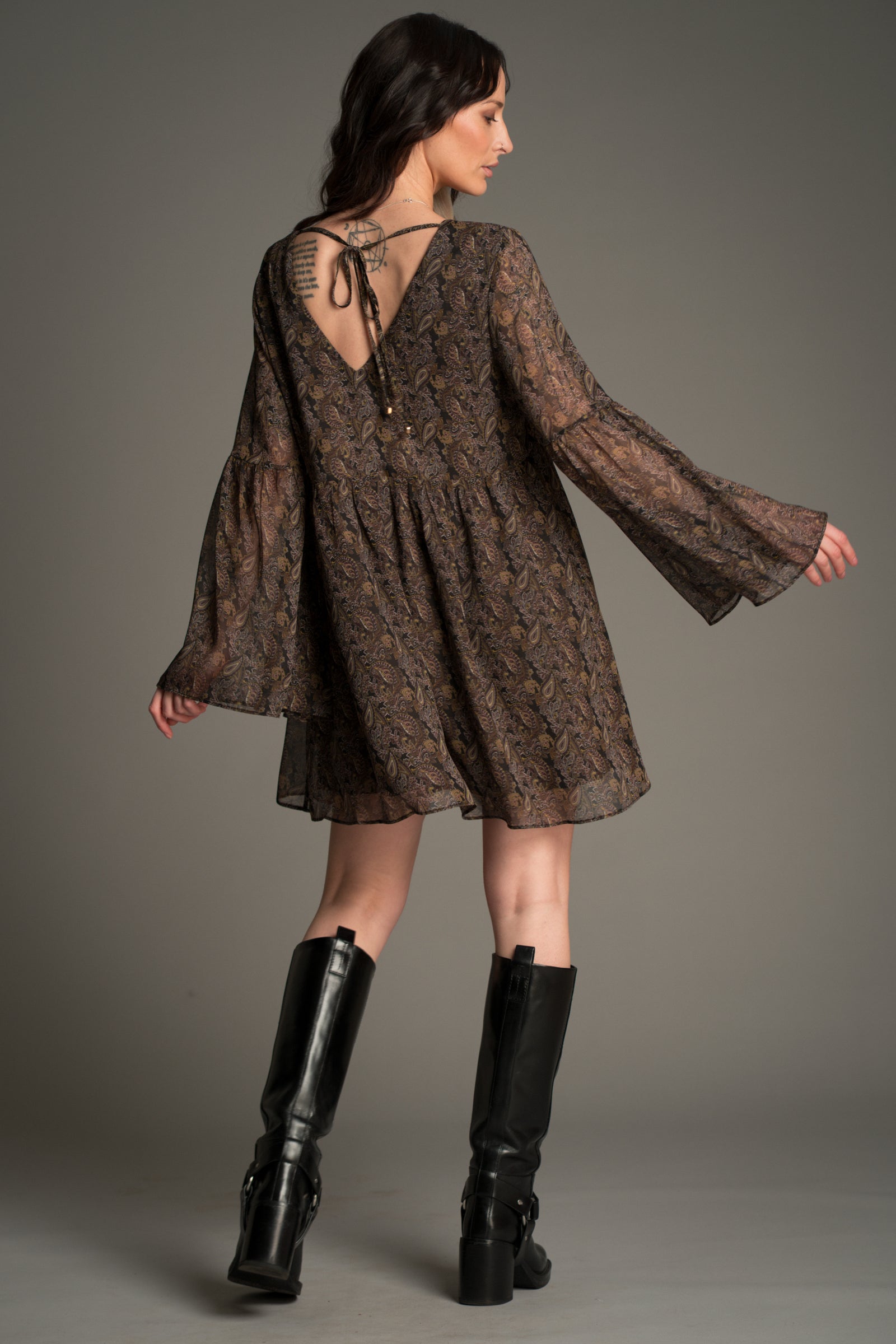 Victoria wears Little Lies Cinnamon Girl Paisley Chiffon Smock Dress shown from the back, featuring a deep V‑back with tie detail, sheer paisley chiffon fabric, flared sleeves, and a relaxed flowing silhouette paired with tall black boots.