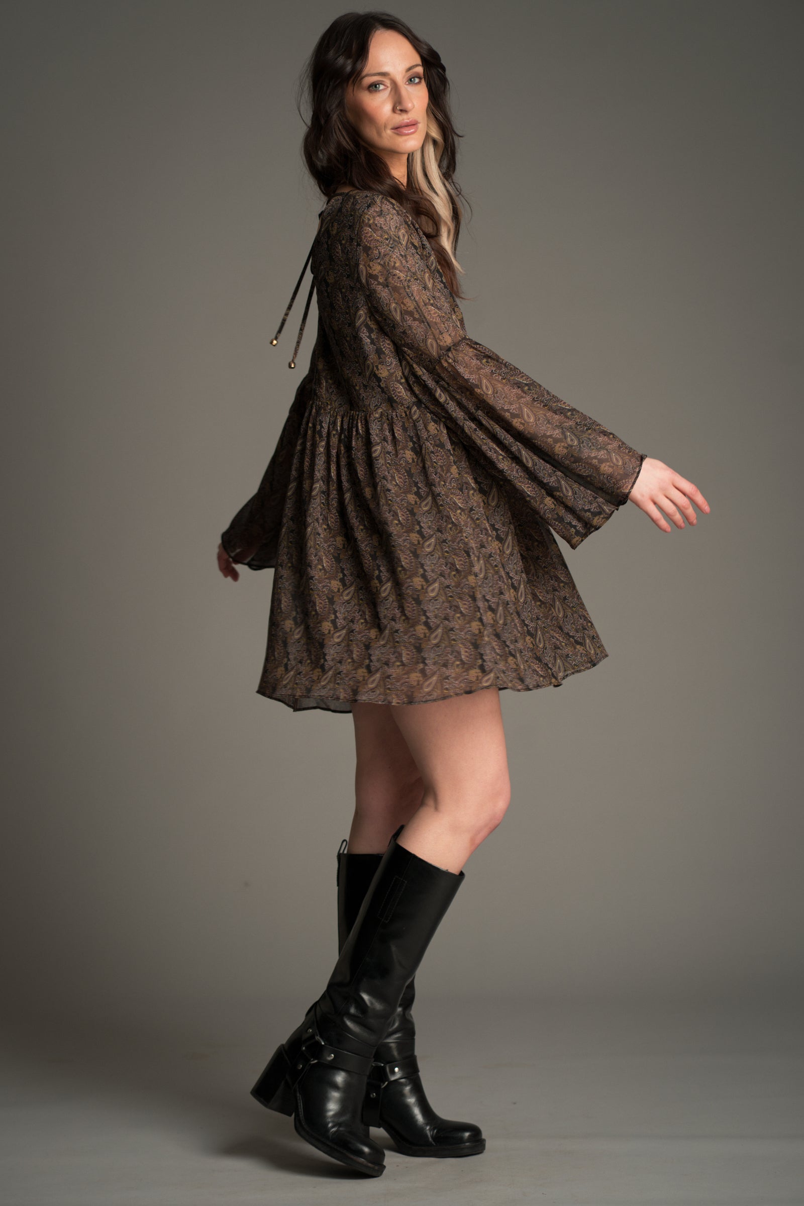 Victoria wears Little Lies Cinnamon Girl Paisley Chiffon Smock Dress shown in a side view, featuring sheer paisley fabric, flared sleeves, a relaxed flowing silhouette, and styled with tall black boots.