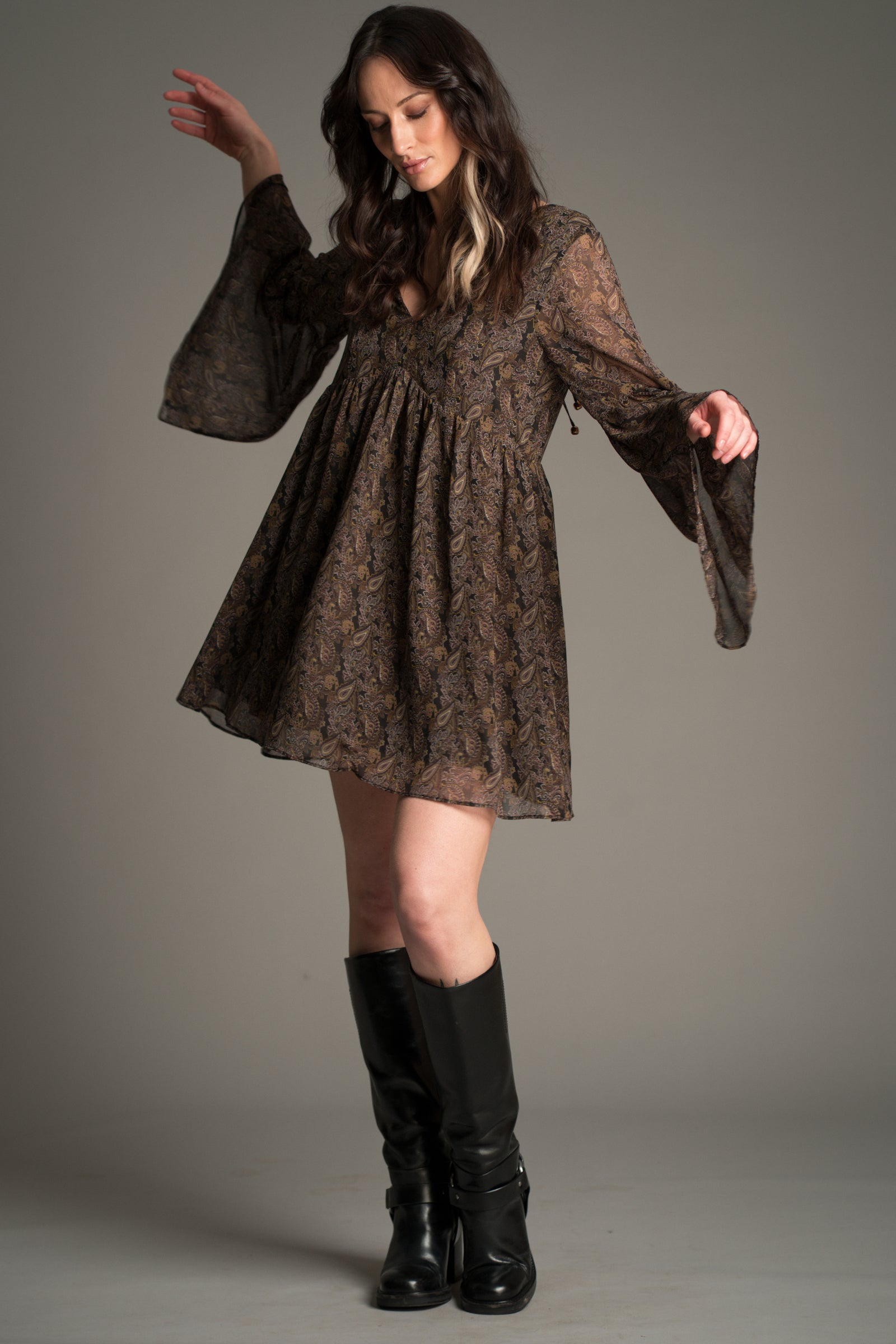 Victoria wears Little Lies Cinnamon Girl Paisley Chiffon Smock Dress with a flowing silhouette, sheer paisley fabric, flared sleeves, and a soft gathered waist, styled with tall black boots.