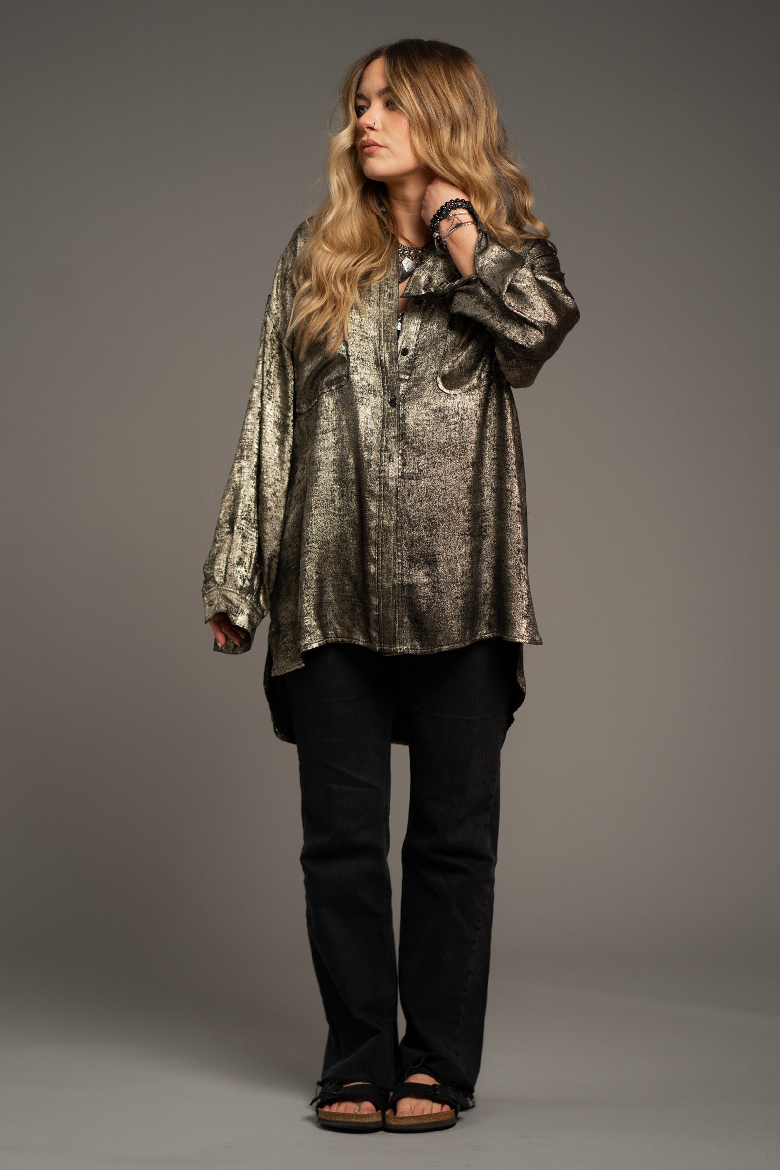 Erin wears Little Lies Luna Metallic Shirt in gold with textured metallic finish, worn in a relaxed oversized style with dark trousers and sandals.