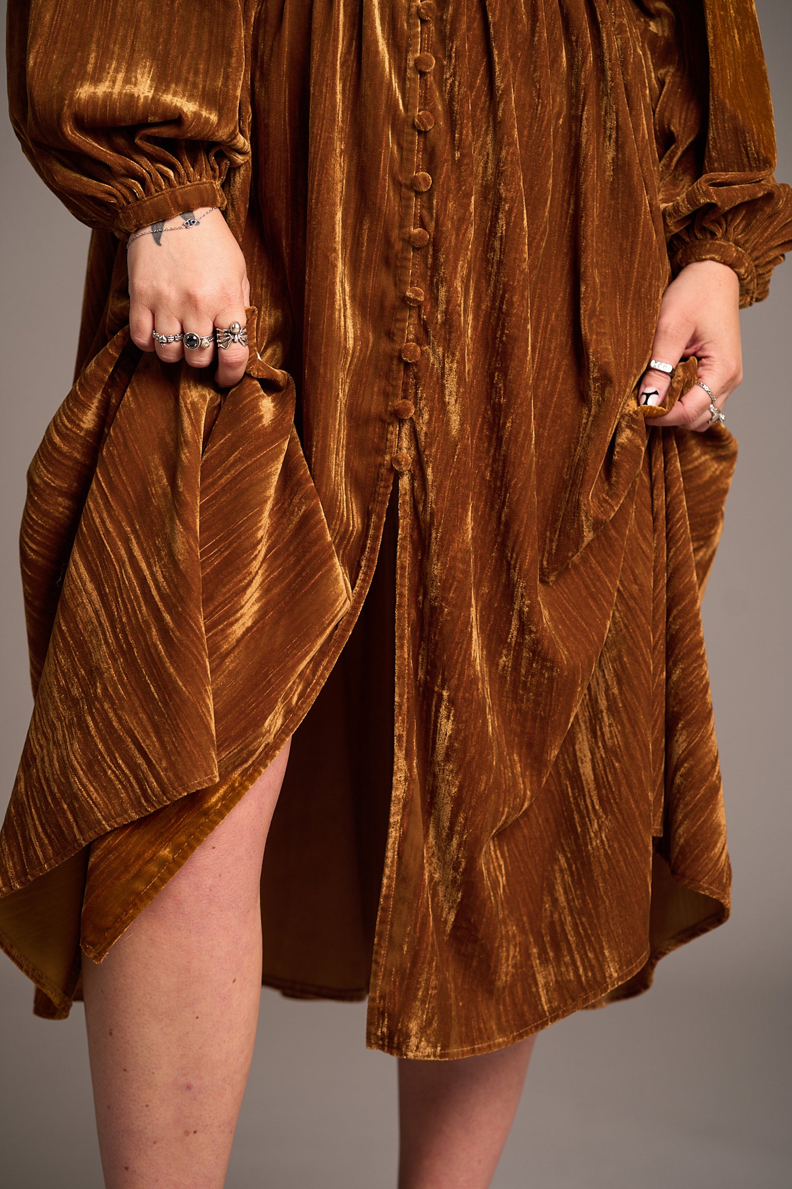 Close-up of Erin wearing Dazed Copper Velvet Midi Dress with button front, long sleeves, lifted slightly to reveal legs and accessorized with statement rings