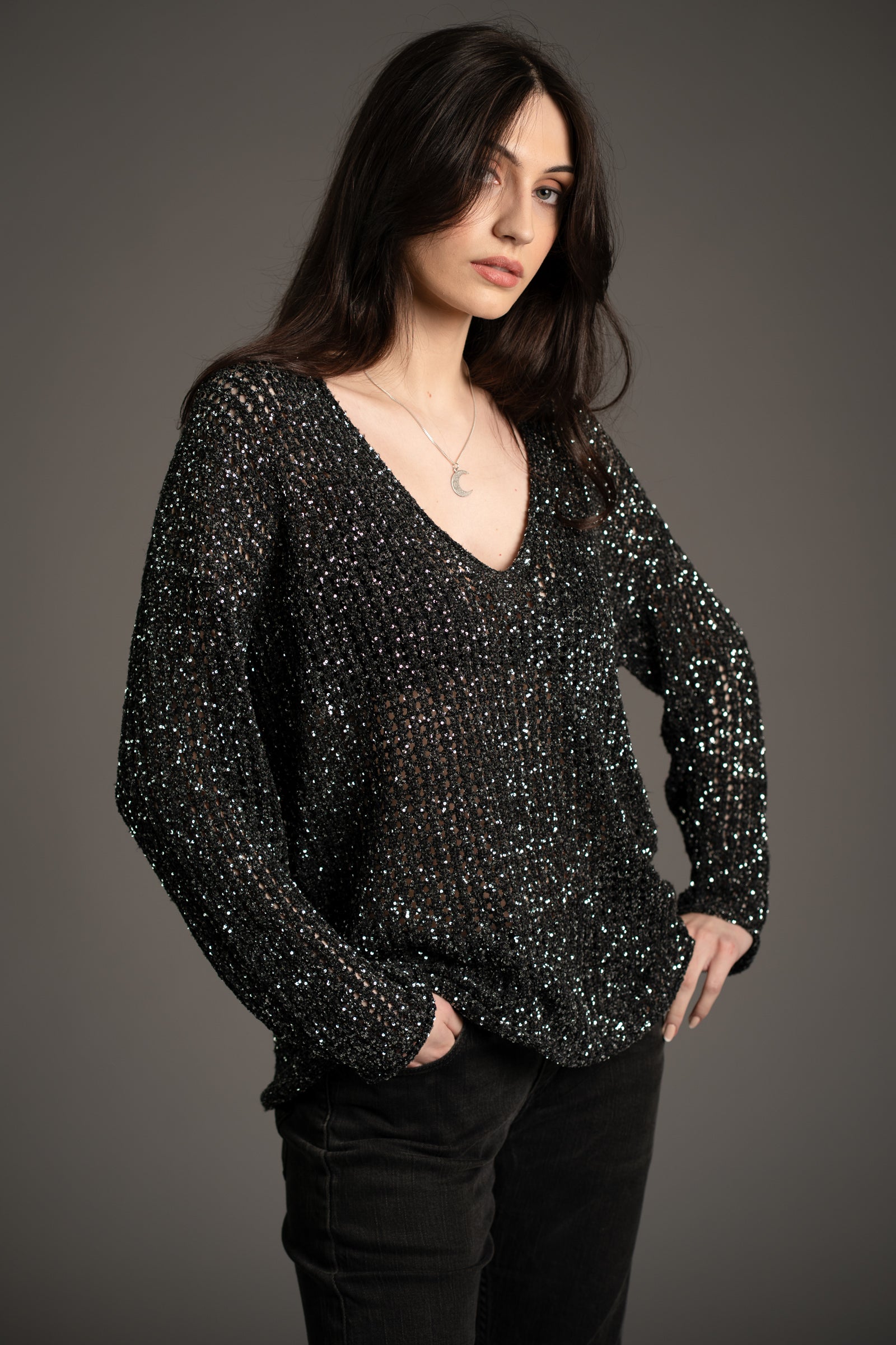 Andie models Little Lies Dazed & Confused sequin knit sweater with deep V-neckline, shimmering sequin detail, styled with dark jeans in studio setting