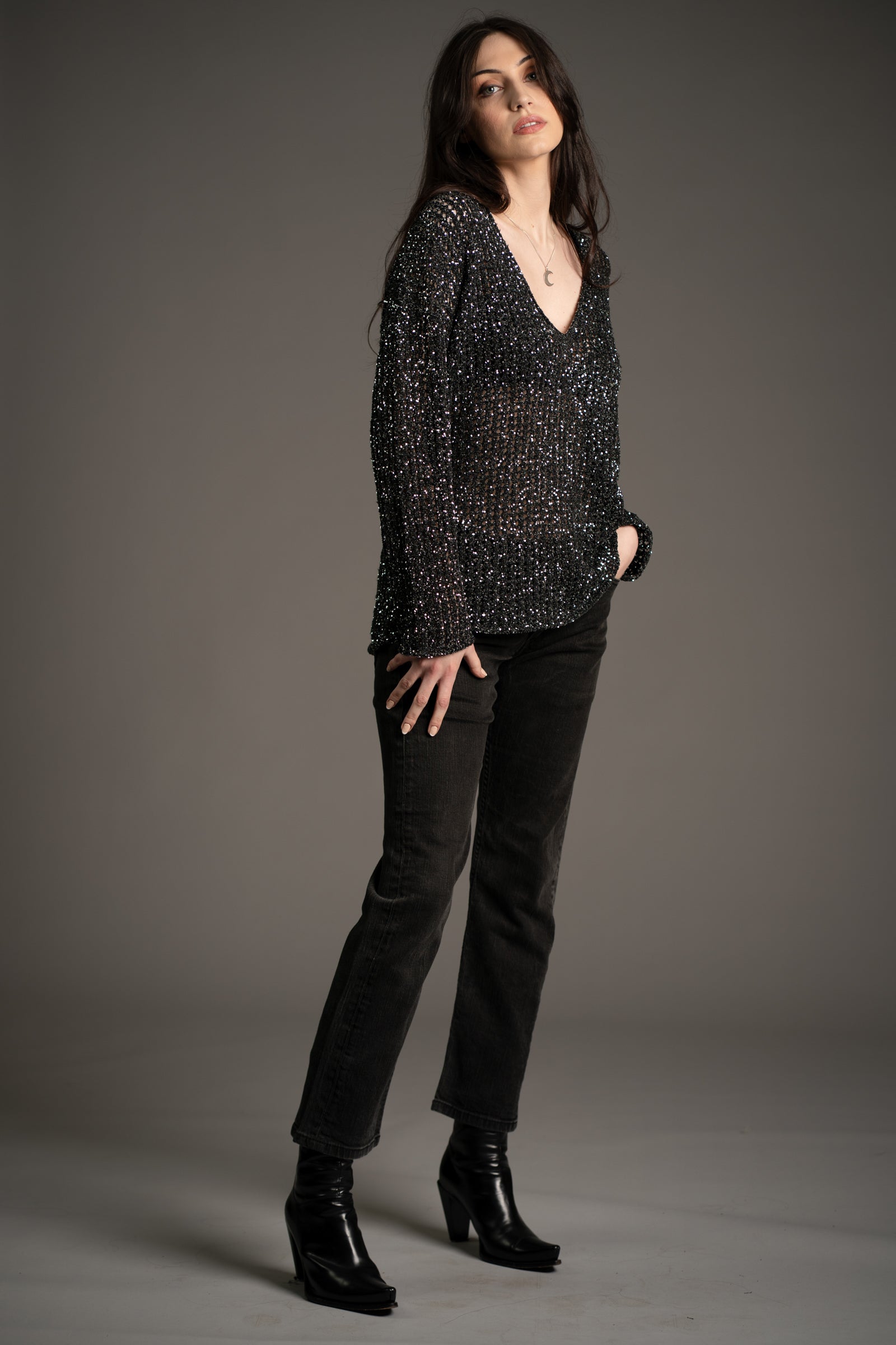 Andie models Little Lies Dazed & Confused sequin knit sweater with deep V-neckline, shimmering sequin detail, styled with straight-leg black jeans and heeled ankle boots in studio setting