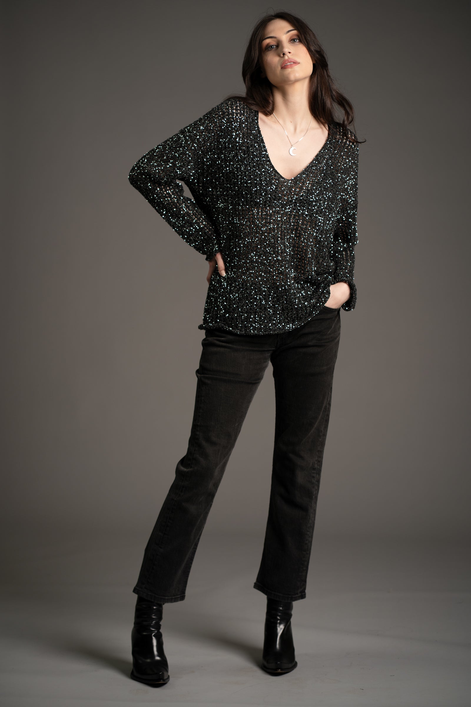 Andie models Little Lies Dazed & Confused sequin knit sweater with deep V-neckline, shimmering sequin detail, styled with straight-leg black jeans and heeled ankle boots in studio setting