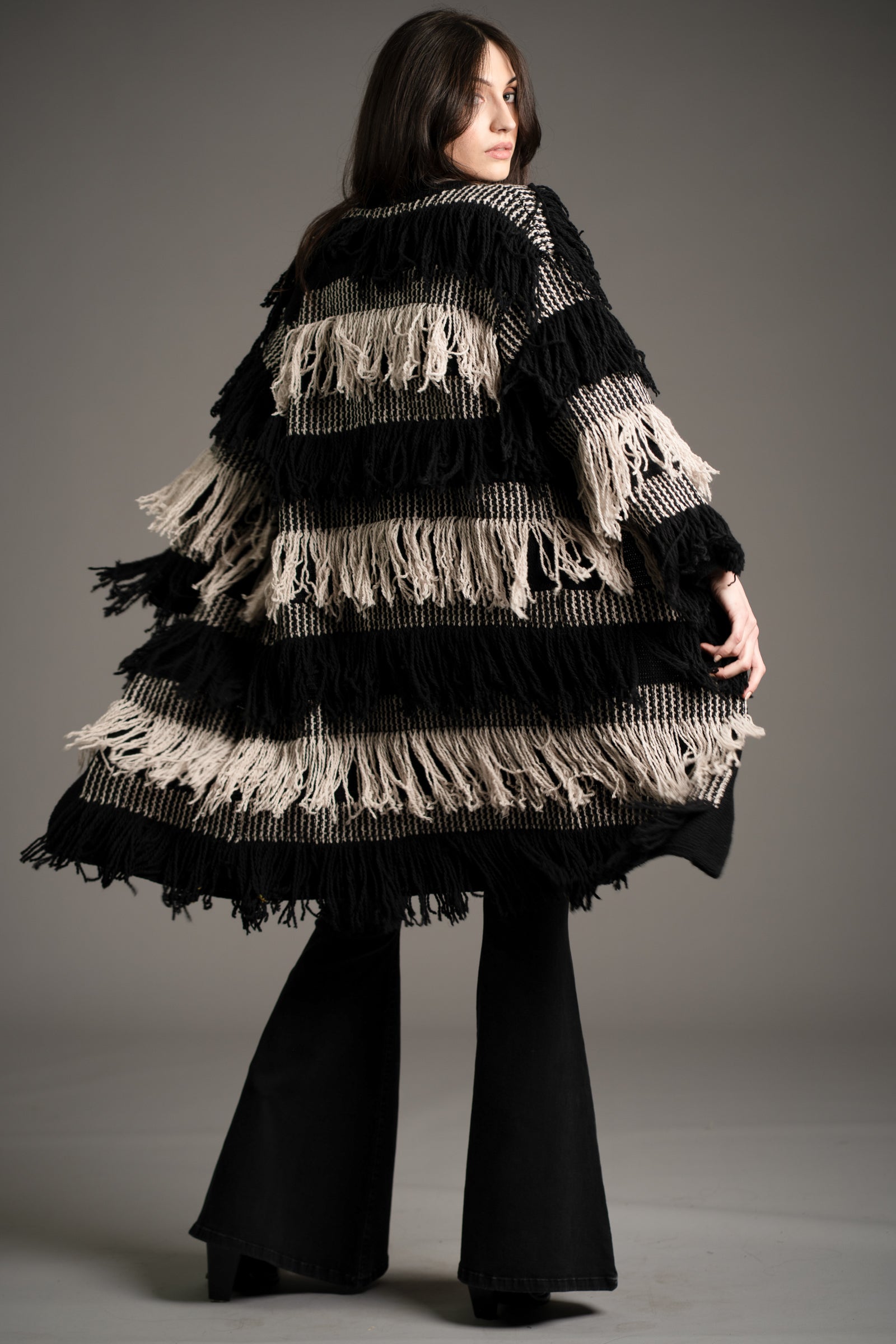 Back view of Andie modelling the Little Lies Desperado Shaggy Cardigan in black and cream fringe design, styled with black flared jeans, studio setting