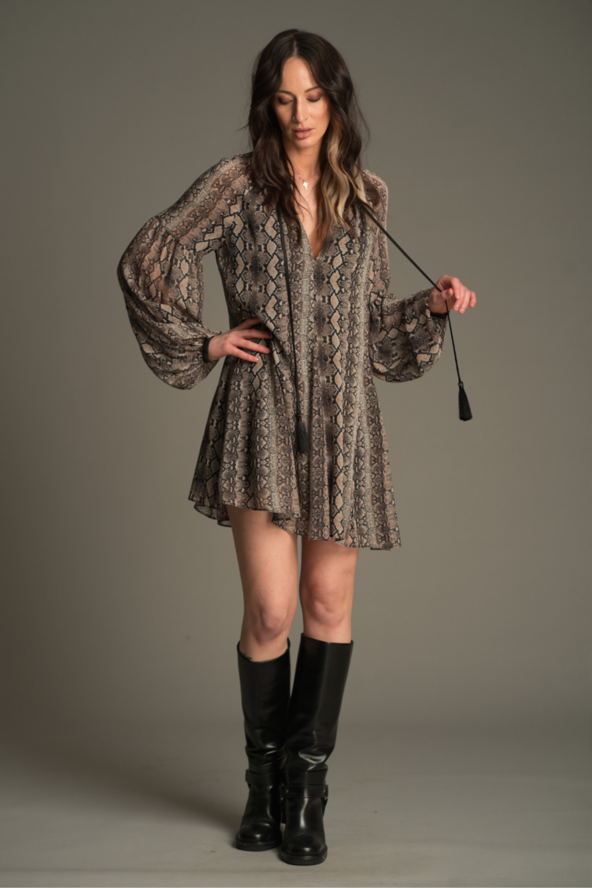 Model wearing the Little Lies Seeker Snake Mini Dress featuring a flowing snake‑print fabric, deep V‑neckline, and long voluminous sleeves, styled with knee‑high black boots.