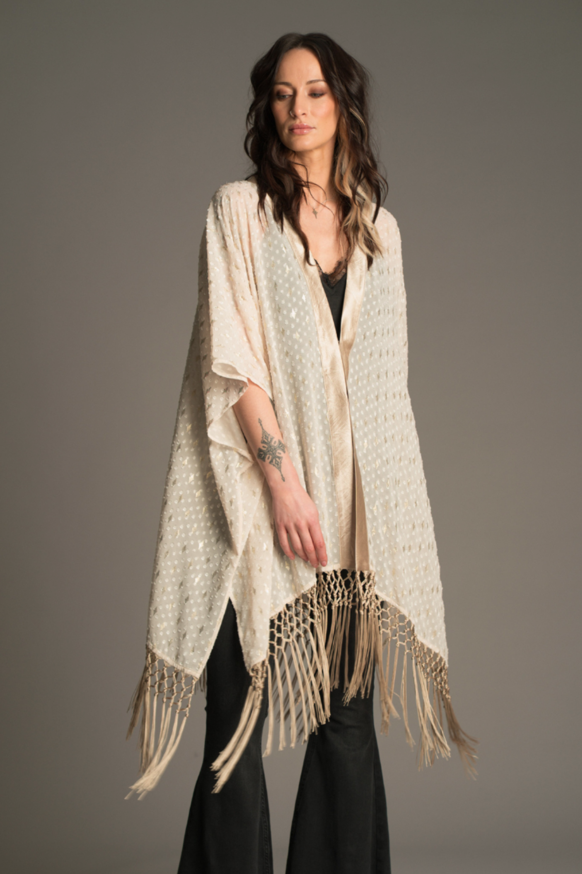 model wearing the Little Lies Songbird Ivory Kimono featuring a soft ivory draped fabric with subtle textured detailing and long fringe trim, styled over a black satin cami and flared denim trousers.