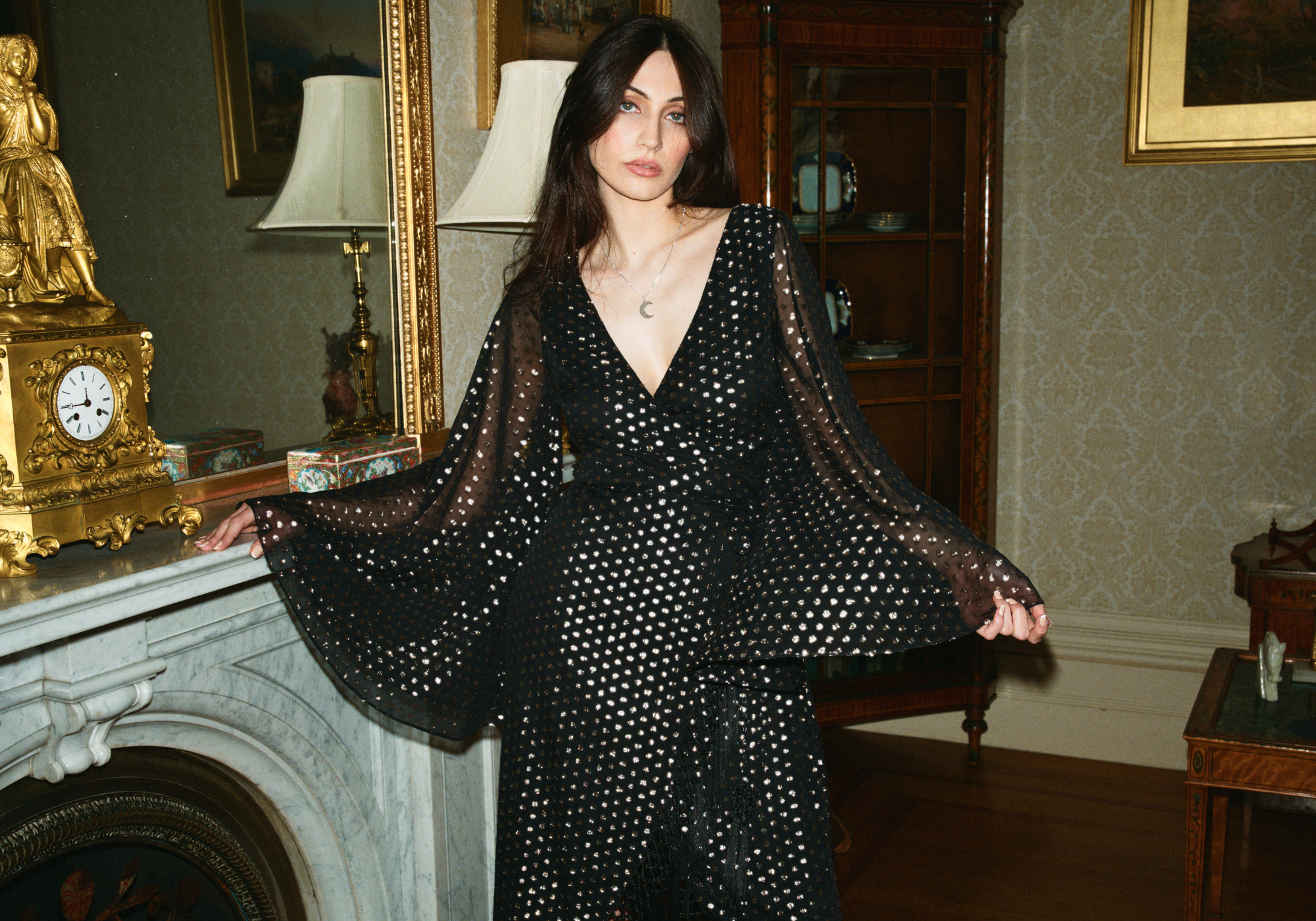 Andie models Little Lies Cosmic Dancer Metallic Wrap Dress in black featuring shimmering metallic polka dot pattern, dramatic flared sleeves, wrap silhouette, in an elegant vintage interior