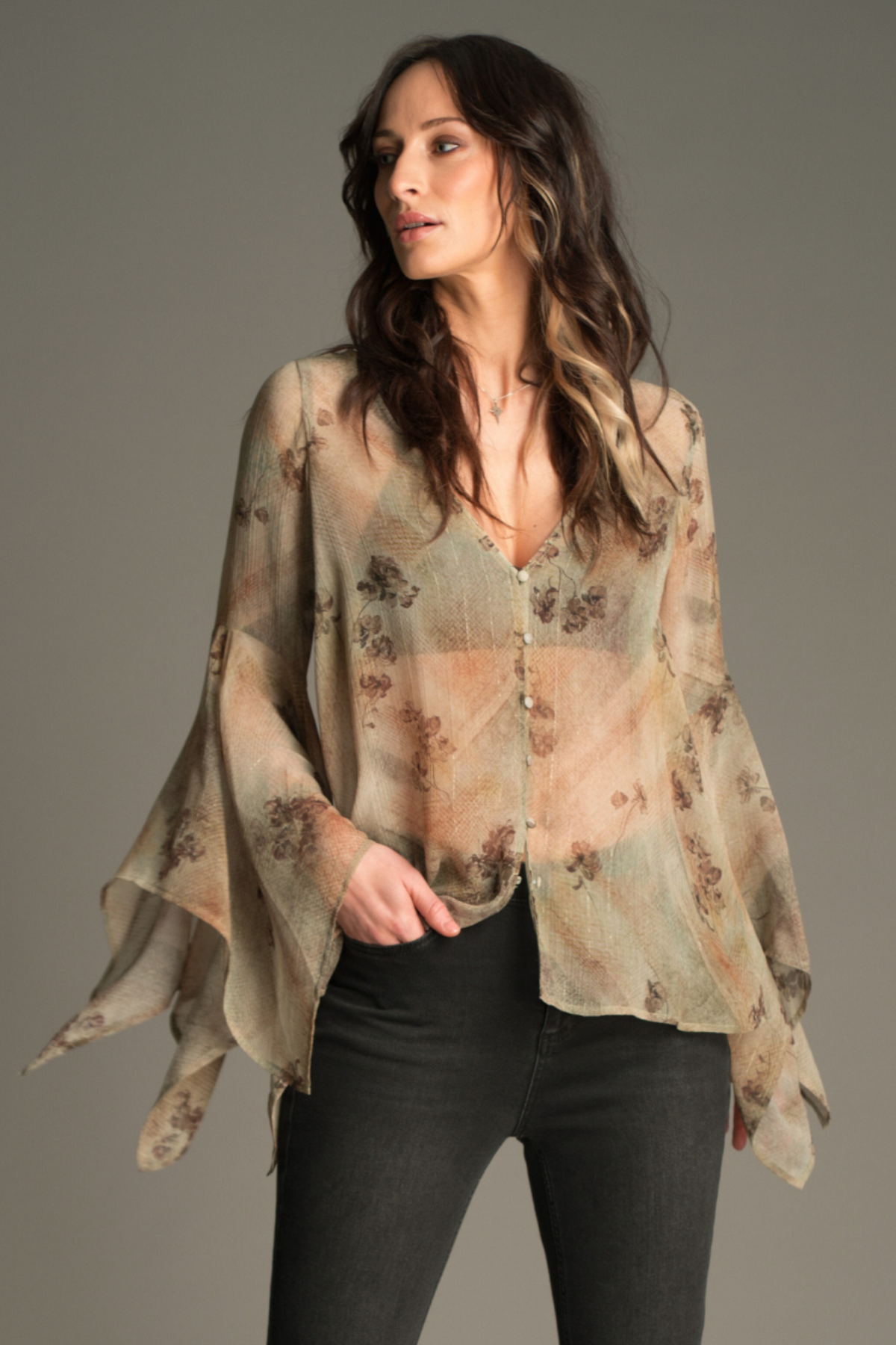 Model wearing the Little Lies Karma Sage Blouse in soft sage floral print with flowing asymmetric sleeves, styled with denim flared trousers.