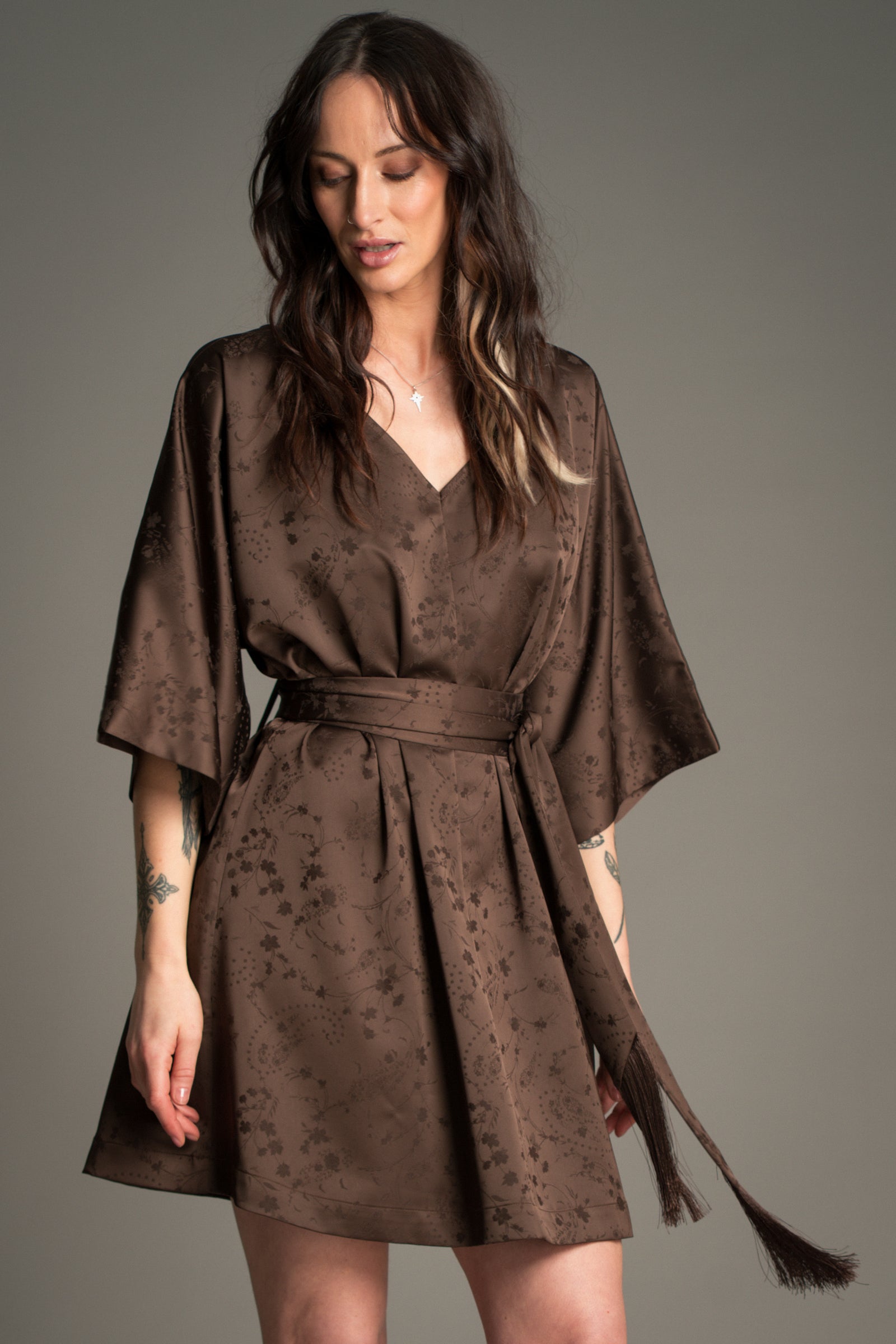 Victoria wears Little Lies Evermore Espresso Batwing Mini Dress featuring wide batwing sleeves, a V‑neckline, textured espresso‑brown fabric, and a long tassel waist tie.