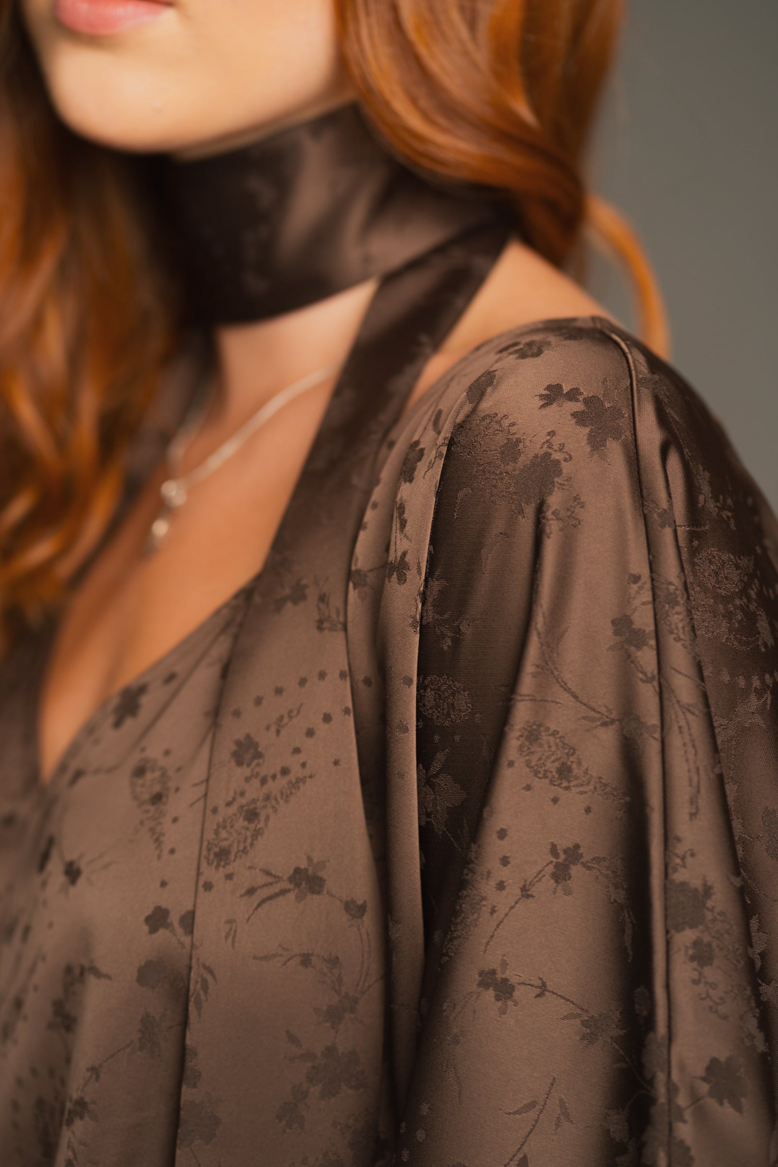 Close-up of a Anna wearing the Evemore Espresso Dress in a brown floral-patterned garment with a blurred background