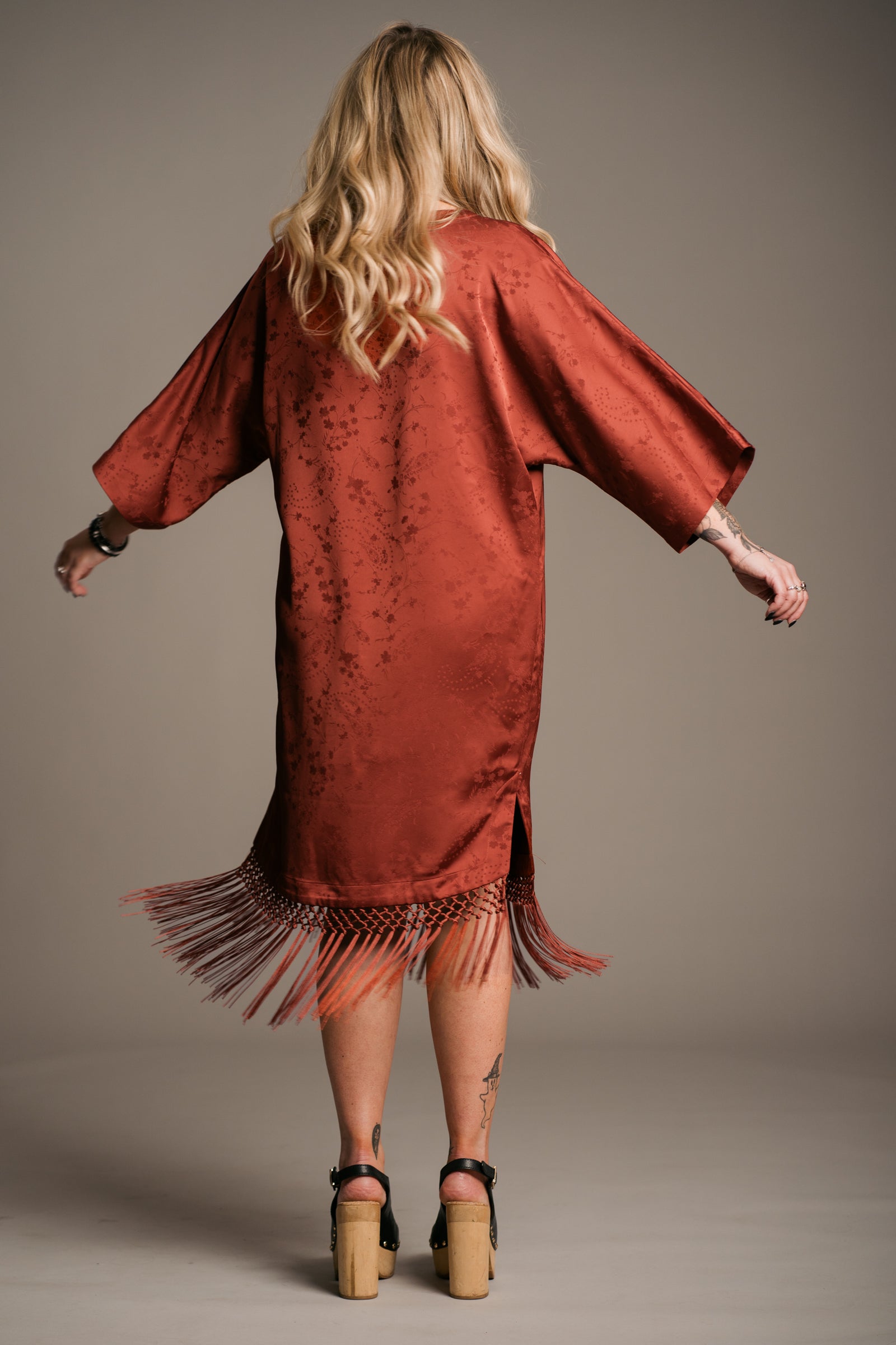 Our Model, Erin, posing against a neutral backdrop, wearing the Little Lies Florence rust-colored fringe satin kimono dress with three-quarter sleeves and fringing along the hem. Paired with black wooden platform clogs, the outfit features a relaxed bohemian style. The model has long, wavy blonde hair and a visible tattoos.