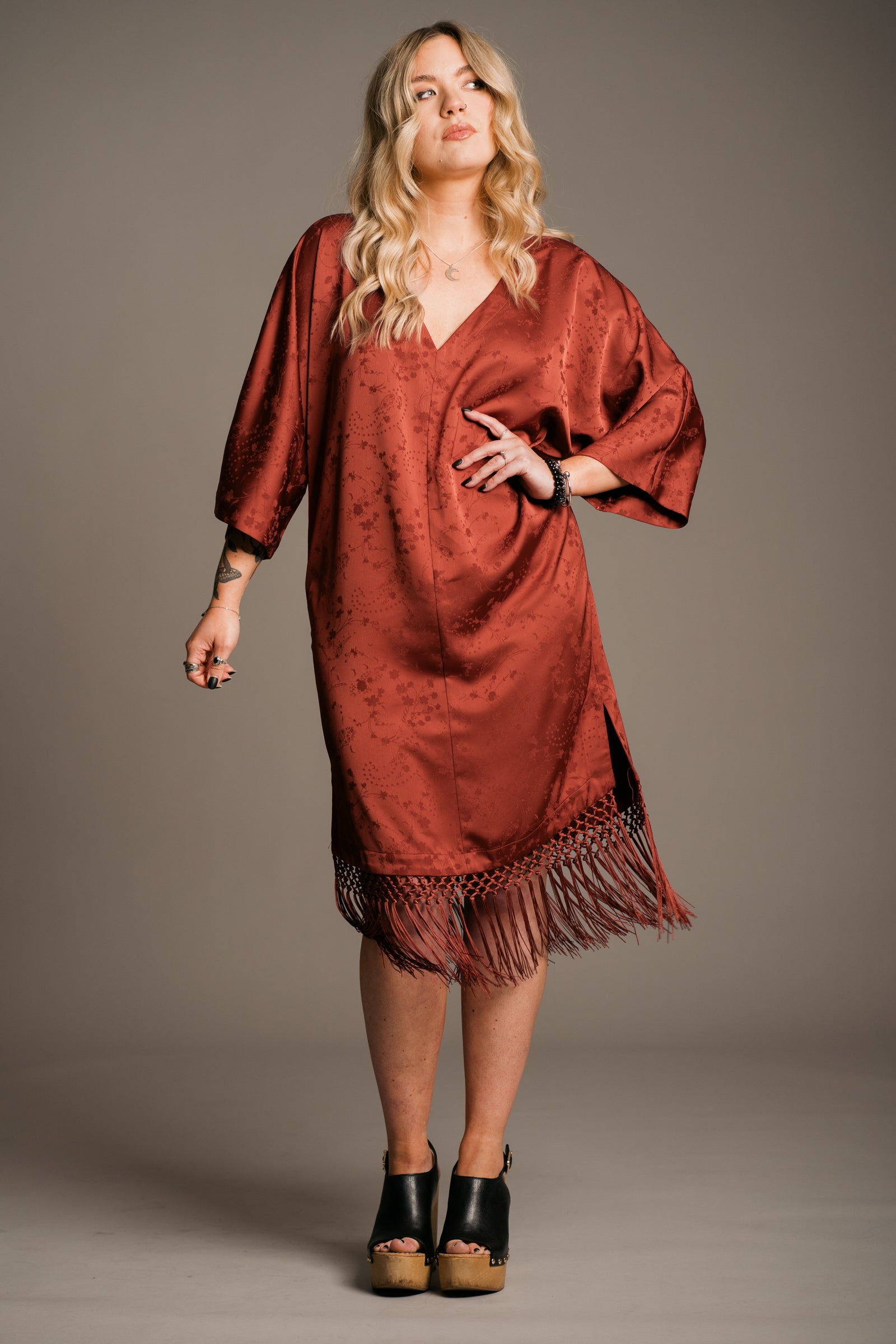 Our Model, Erin, posing against a neutral backdrop, wearing the Little Lies Florence rust-colored fringe satin kimono dress with three-quarter sleeves and a V-neckline. Paired with black wooden platform clogs, and the Little Lies Crescent sterling silver necklace, the outfit features a relaxed bohemian style. The model has long, wavy blonde hair and a visible tattoo on the left forearm.