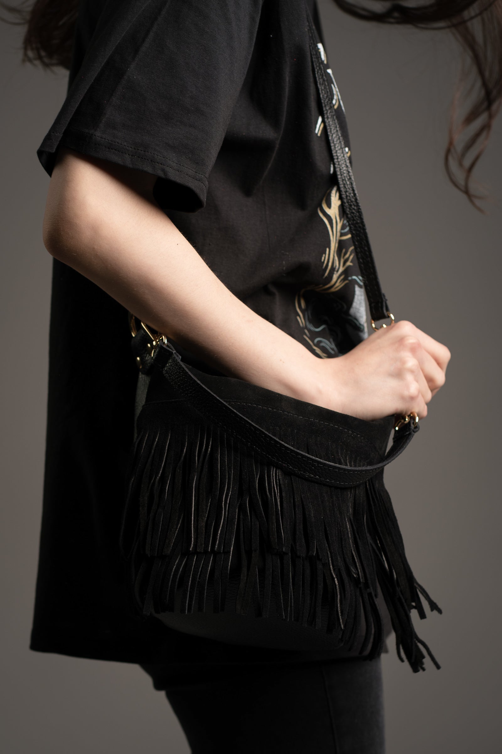 Model holding Gemini Genuine Leather Fringed Bucket Bag in black with gold hardware, dual straps and layered fringe detail by Little Lies in studio setting
