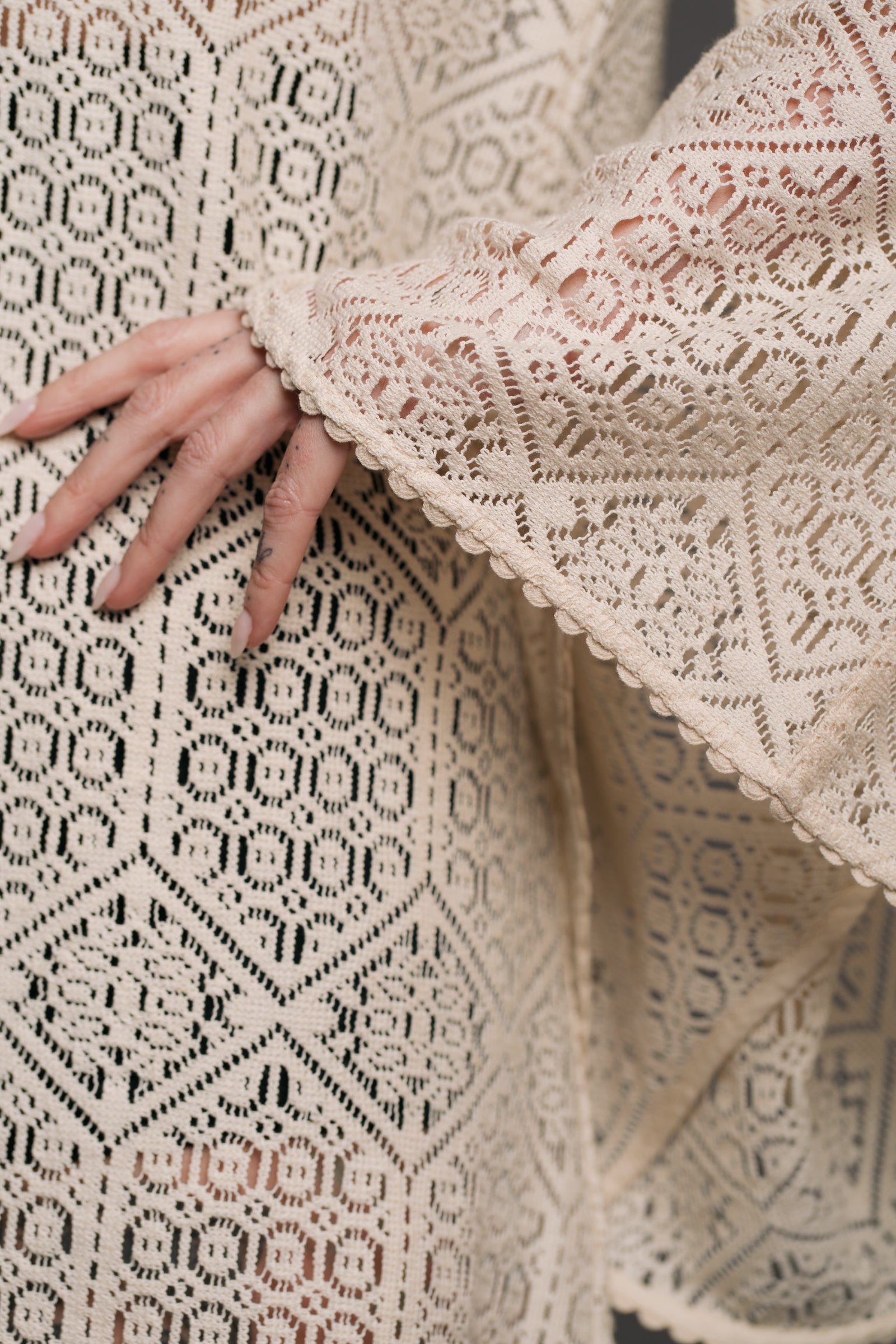 Our model, Bex, standing against a neutral backdrop in the studio wearing the Little Lies Goldie Ivory Crochet Bell Sleeved Mini Dress, featuring intricate crochet detailing, flowing bell sleeves, and a flattering mini silhouette. Close up shot showing the details within the ivory crochet style fabric - effortless bohemian elegance.
