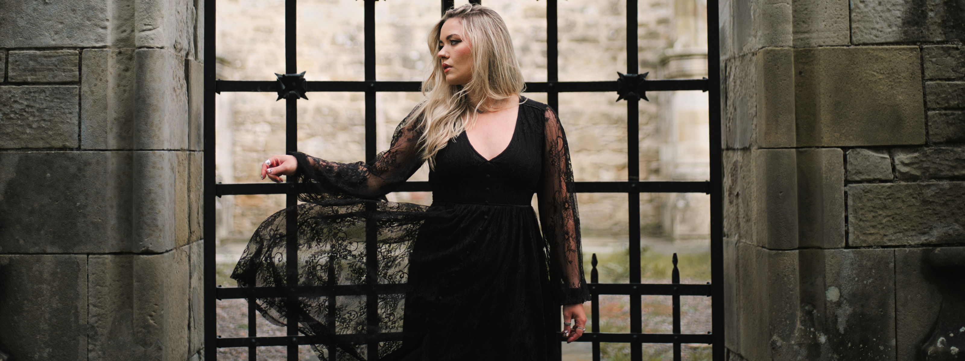 Close up of Erin modelling the Little Lies Dazed Black Lace Midi Dress with sheer long sleeves and flared hem, in front of gothic stone arch and wrought iron gate