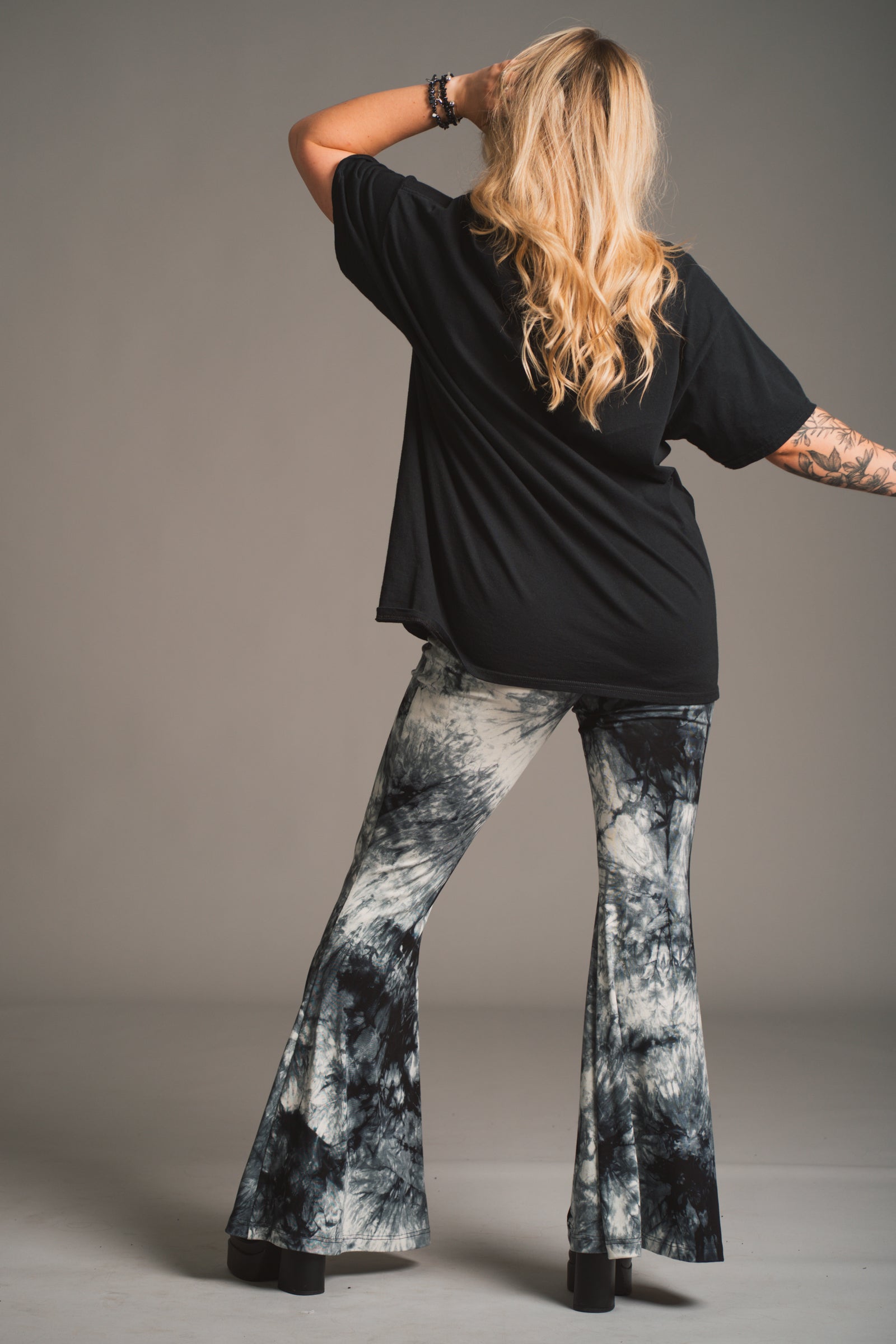 Back view of Erin modelling Little Lies Hendrix tie dye flared trousers styled with loose black tee in studio setting