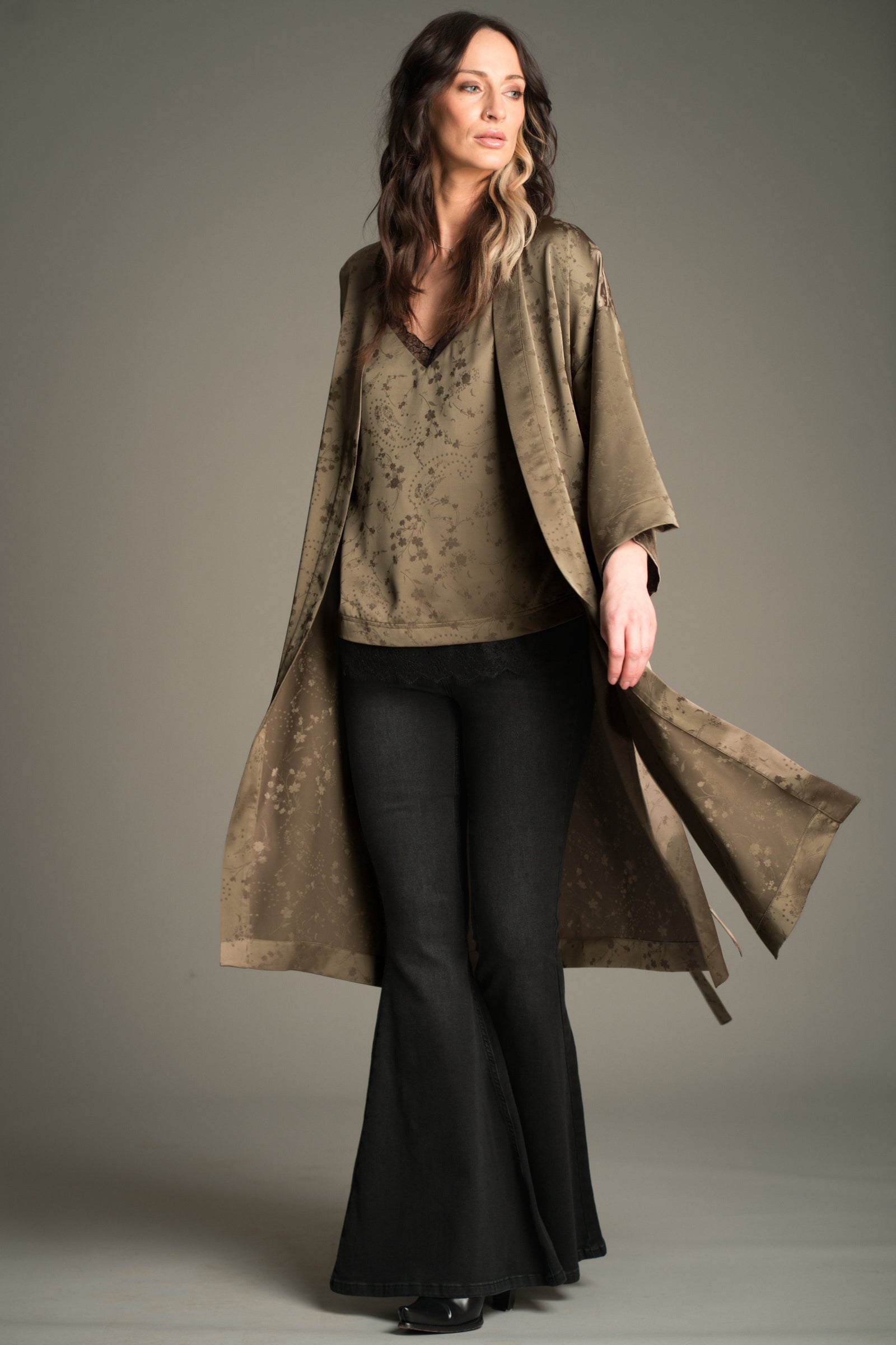 Front view of Victoria wearing the Little Lies Iris Satin Kimono in olive satin with a subtle floral jacquard pattern, layered over a matching Iris Satin cami and styled with denim flared trousers.