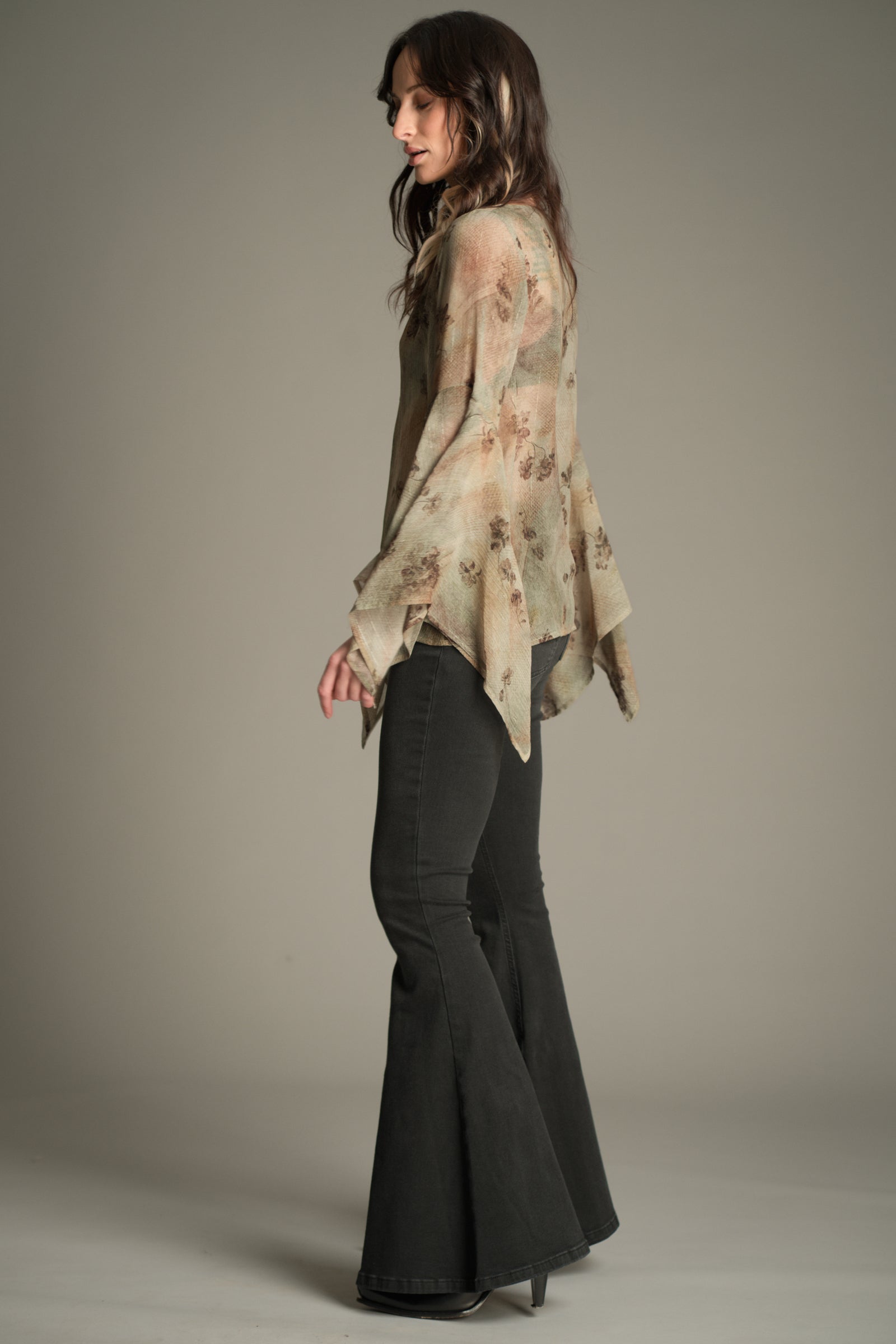 Side‑back view of Victoria wearing the Little Lies Karma Sage Blouse in soft sage floral print with flowing asymmetric flared sleeves, styled with denim flared trousers.