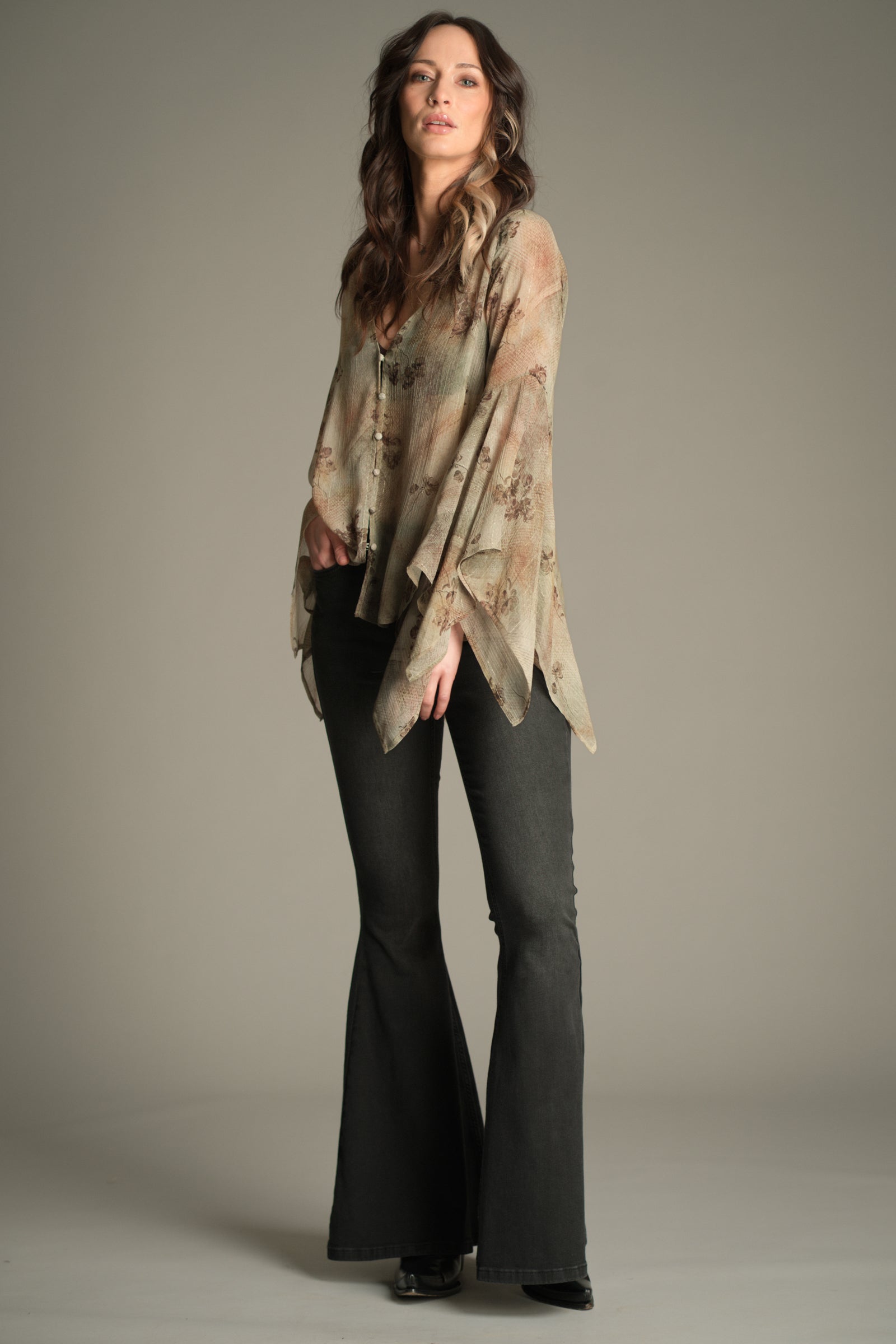 Full‑length view of Victoria wearing the Little Lies Karma Sage Blouse in soft sage floral print with flowing asymmetric sleeves, styled with denim flared trousers.