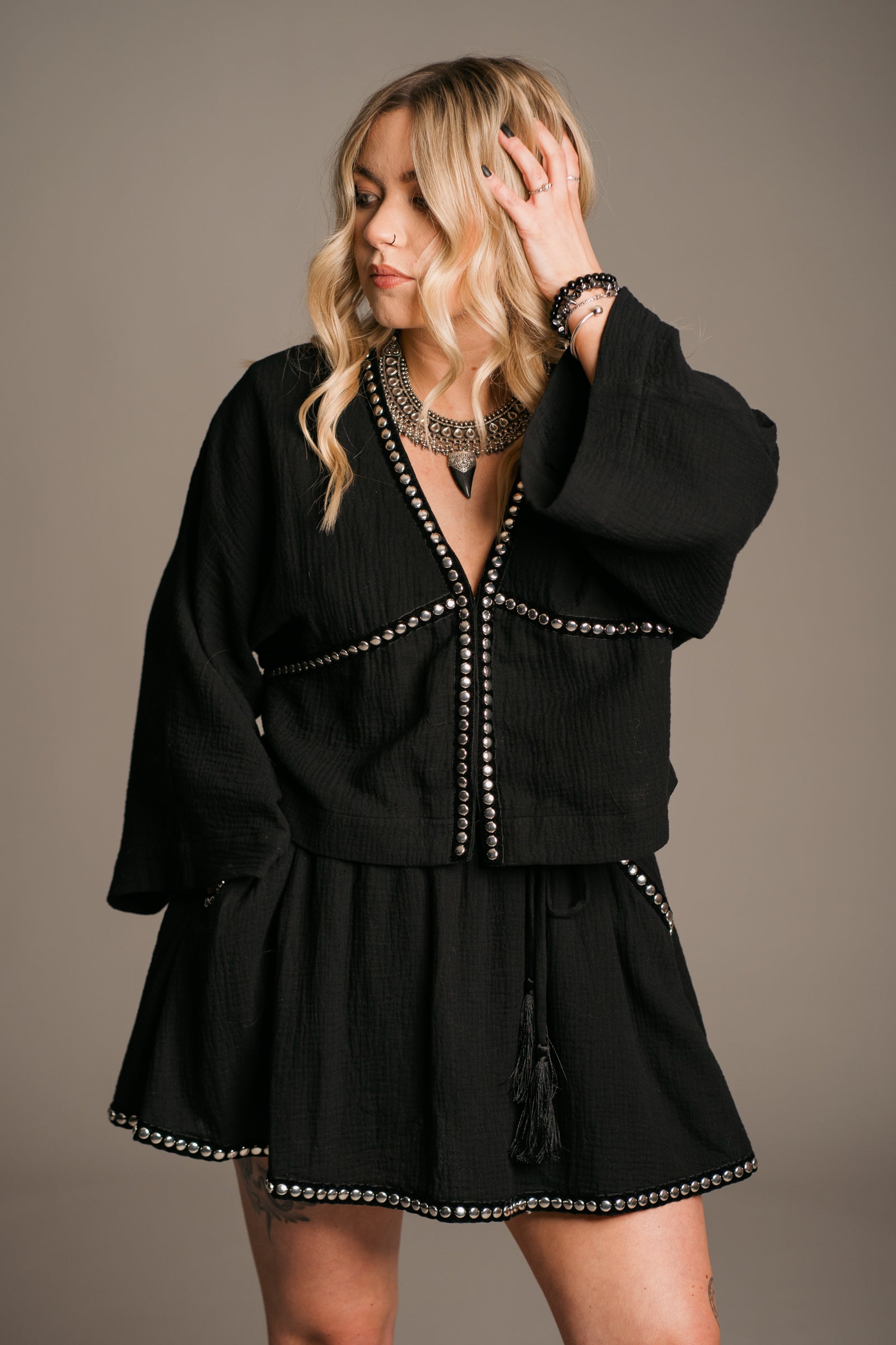 Blonde model Erin wearing the Little Lies Kravitz Black Studded Cheesecloth Co-ord, featuring silver studding along the neckline of the Kravitz Studded Top and detailed studding on the hem and pockets of the Kravitz Studded Skort. The skort is styled with a cord tie waist belt and functional pockets. Paired with a Gypsy Silver Tooth necklace, the look embodies effortless Rock 'n' Roll style