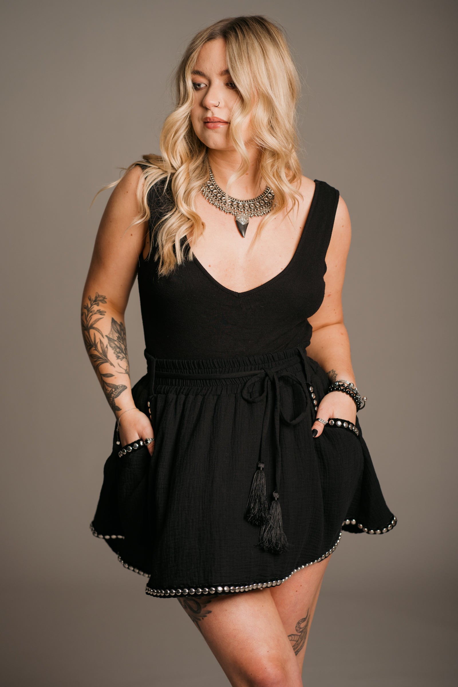 Blonde model Erin wearing the Little Lies Kravitz Black Studded Cheesecloth Skort, featuring silver stud details on the hem and pockets of the Kravitz Studded Skort. The skort has an elasticated chirred waist with a cord tie belt and functional pockets. Paired with a simple black vest top and a Gypsy Silver Tooth necklace, the look embodies effortless Rock 'n' Roll style