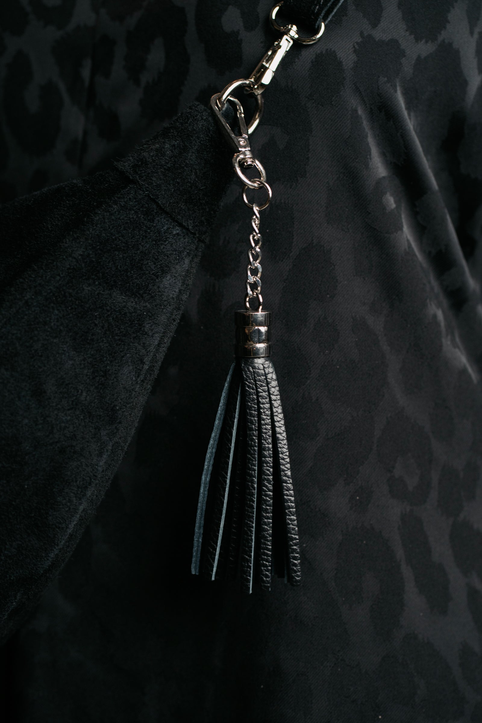 A black genuine leather tassel bag charm / keyring hanging on a dark background.