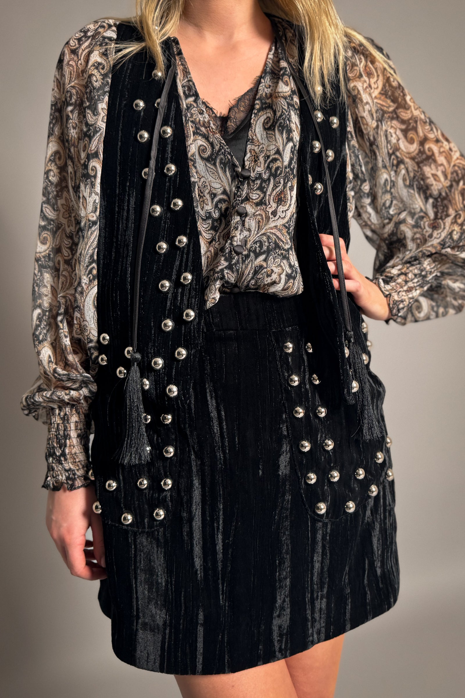 Model wearing Little Lies Lennon black studded velvet mini skirt styled with matching waistcoat and sheer paisley blouse for relaxed boho outfit