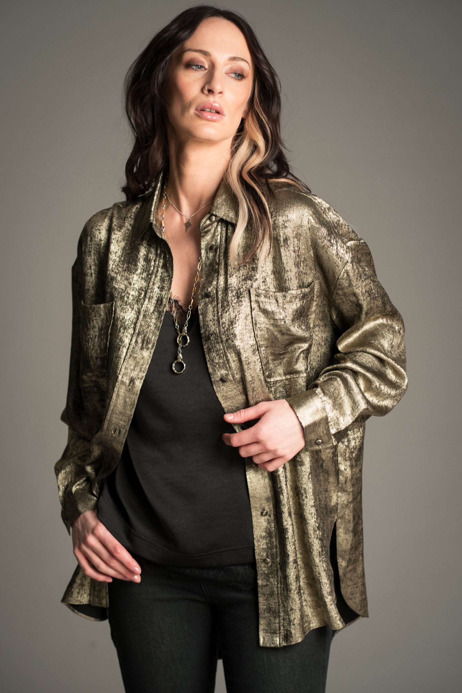 Victoria wears Little Lies Luna Metallic Shirt in gold, front view showing textured metallic finish and relaxed longline fit, styled over black satin cami and denim flares.