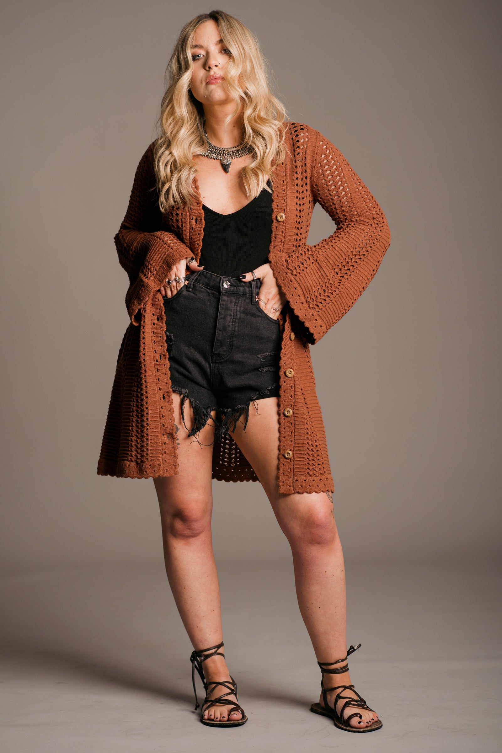 Our model, Erin, stands against a neutral studio backdrop, wearing the Little Lies Marianne Rust Crochet Knit Mini Dress. The dress features intricate crochet detailing, a full button front, a V-neckline, and flared sleeves— styled open also wearing black denim shorts, Little Lies Nightbird Satin top, a Gypsy Silver necklace and black tie up sandals, exuding a relaxed bohemian aesthetic.