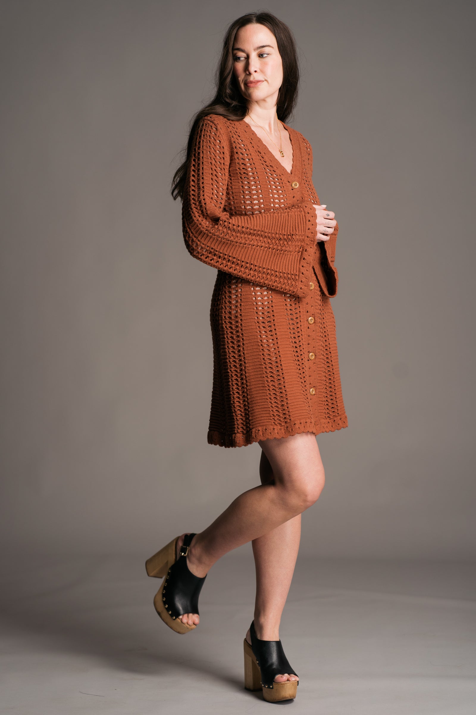 Our model, Bex, stands against a neutral studio backdrop, wearing the Little Lies Marianne Rust Crochet Knit Mini Dress. The dress features intricate crochet detailing, a full button front, a V-neckline, and flared sleeves— styled with black wooden platform clogs, exuding a relaxed bohemian aesthetic.