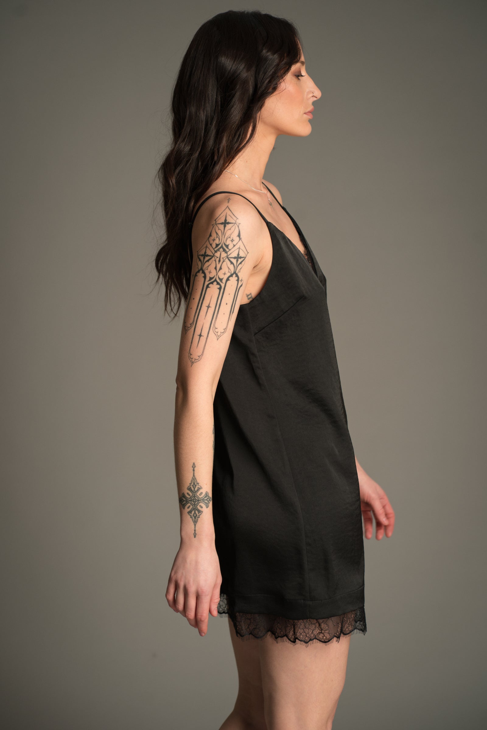 Victoria posing against a grey studio backdrop, wearing the Little Lies Nightbird Black Satin Slip Dress Mini showing the side of the dress with lace trim hem