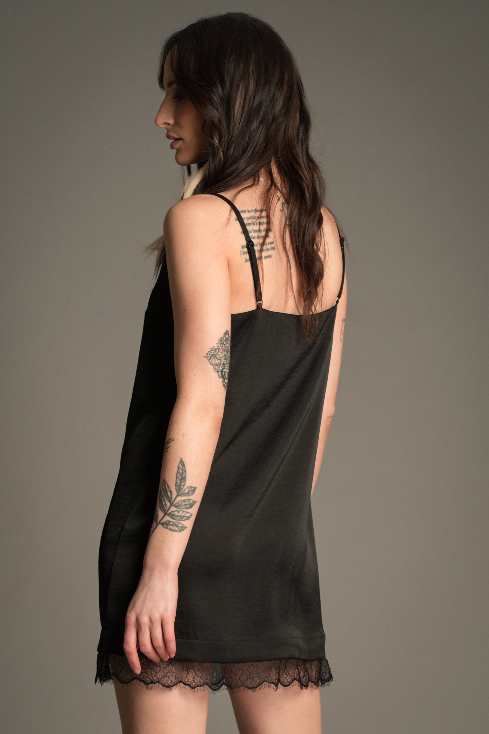Victoria posing against a grey studio backdrop, wearing the Little Lies Nightbird Black Satin Slip Dress Mini showing the back of the dress with lace trim hem and adjustable straps