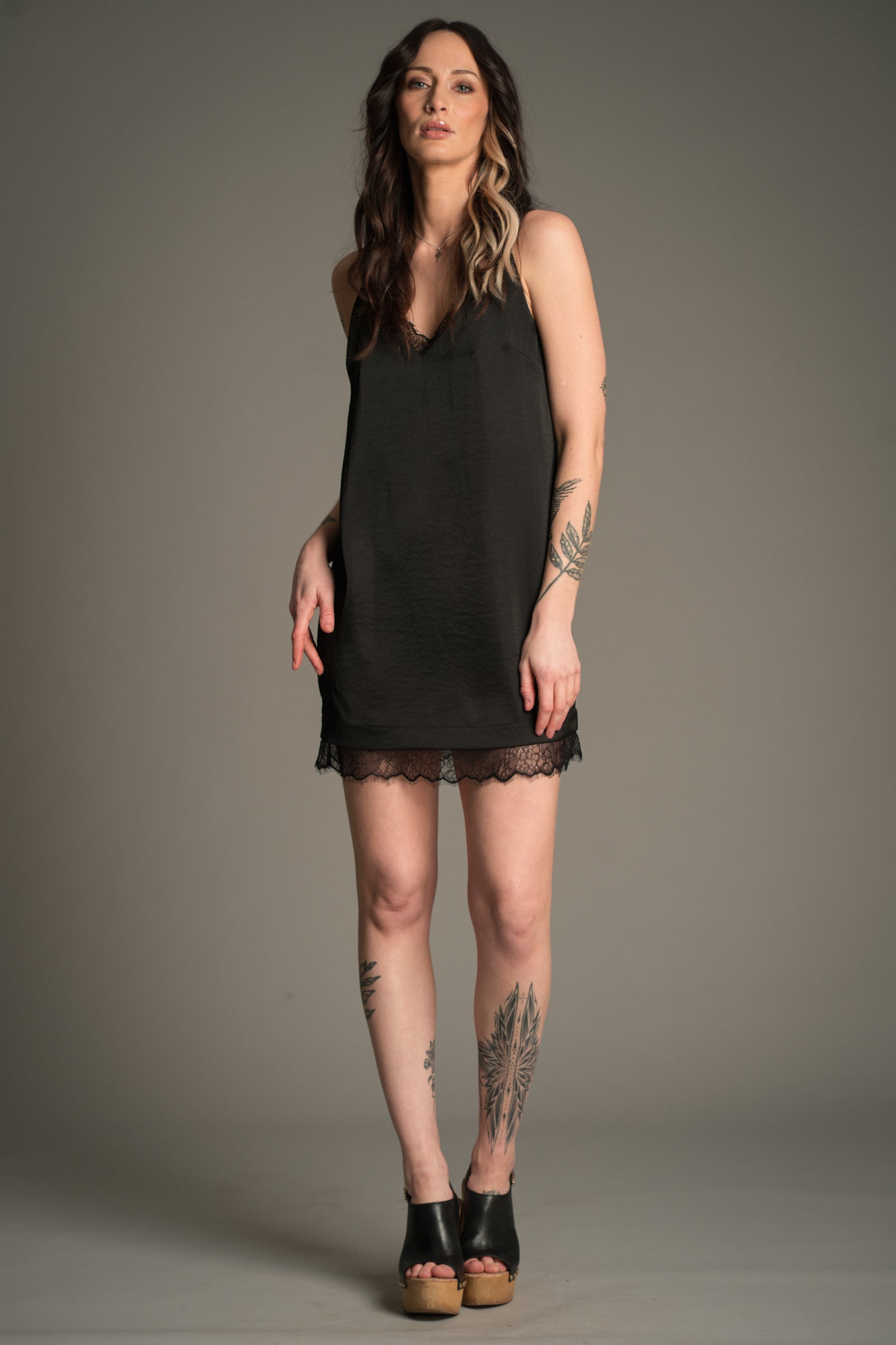 Victoria posing in black Nightbird Satin mini dress with lace trim neckline and hem, styled with wooden heeled clogs