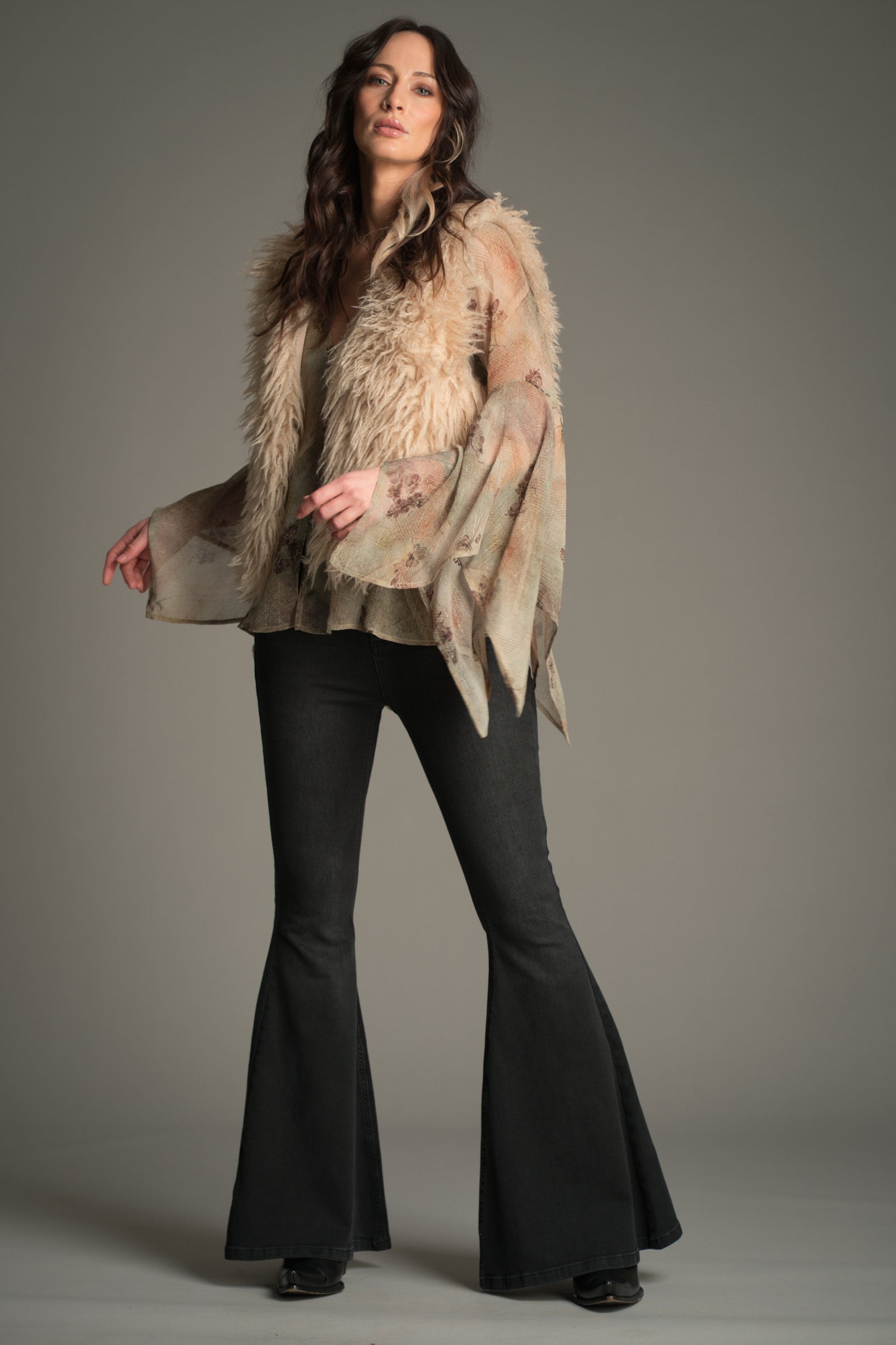 Model wearing the beige Little Lies Penny Lane Faux Mongolian Fur Gilet gilet layered over a Karma sheer floral blouse with wide draped sleeves, paired with Hendrix flared jeans in a studio.