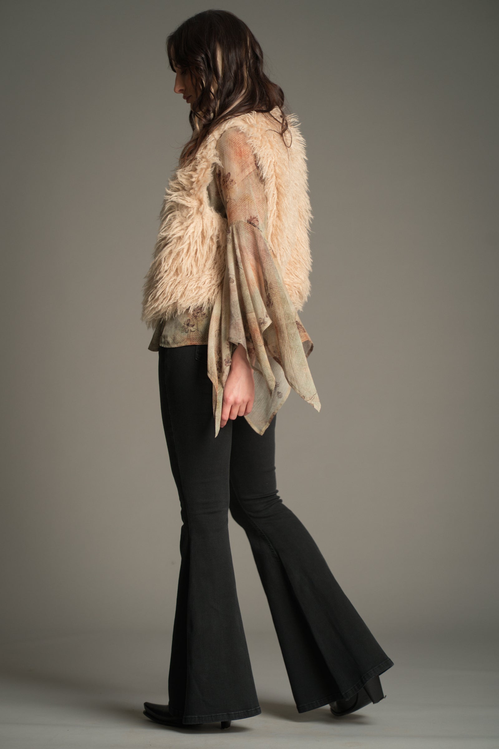Side view of model wearing a beige faux‑fur Little Lies Penny Lane gilet over a Karma sheer floral blouse with draped sleeves, paired with Hendrix flared jeans.