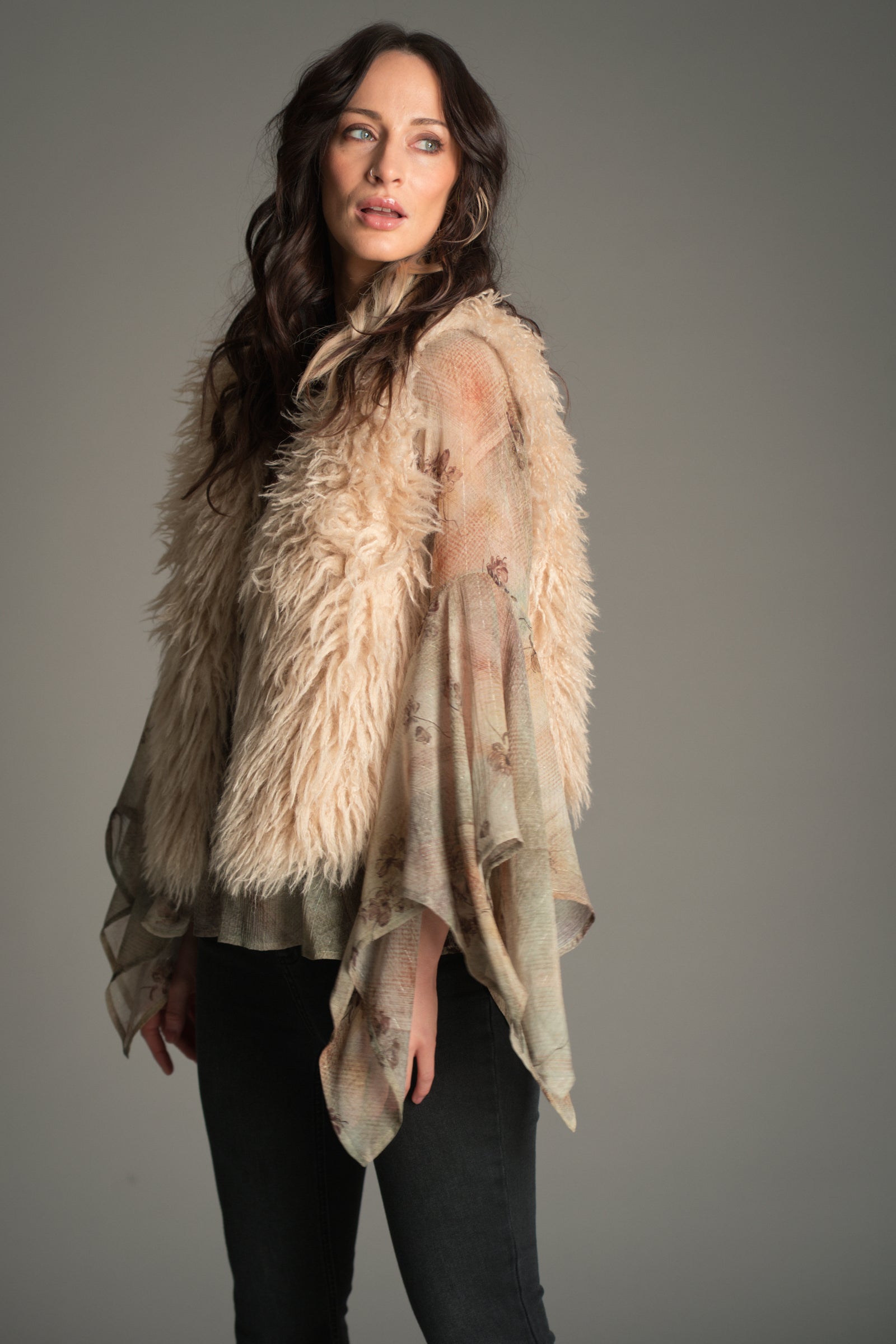 Model wearing the beige Little Lies Penny Lane Faux Mongolian Fur Gilet gilet layered over a Karma sheer floral blouse with wide draped sleeves, paired with Hendrix flared jeans in a studio.