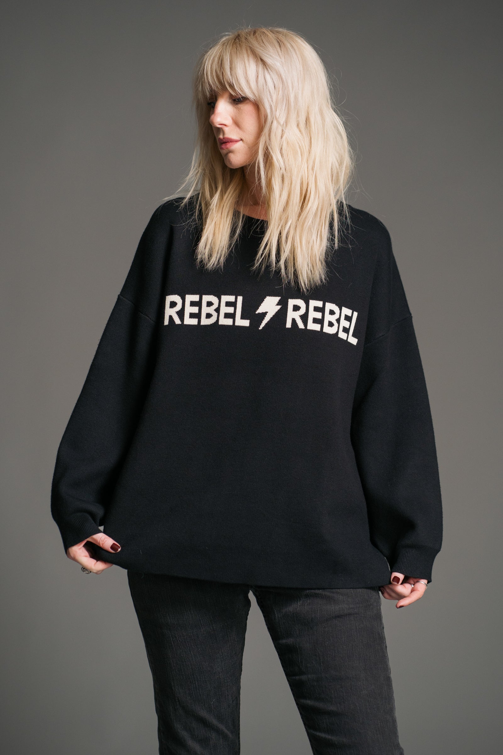 Sara wearing a the Rebel Rebel Black Graphic Jumper with 'REBEL REBEL' ivory text and lightning bolt graphic on a gray background