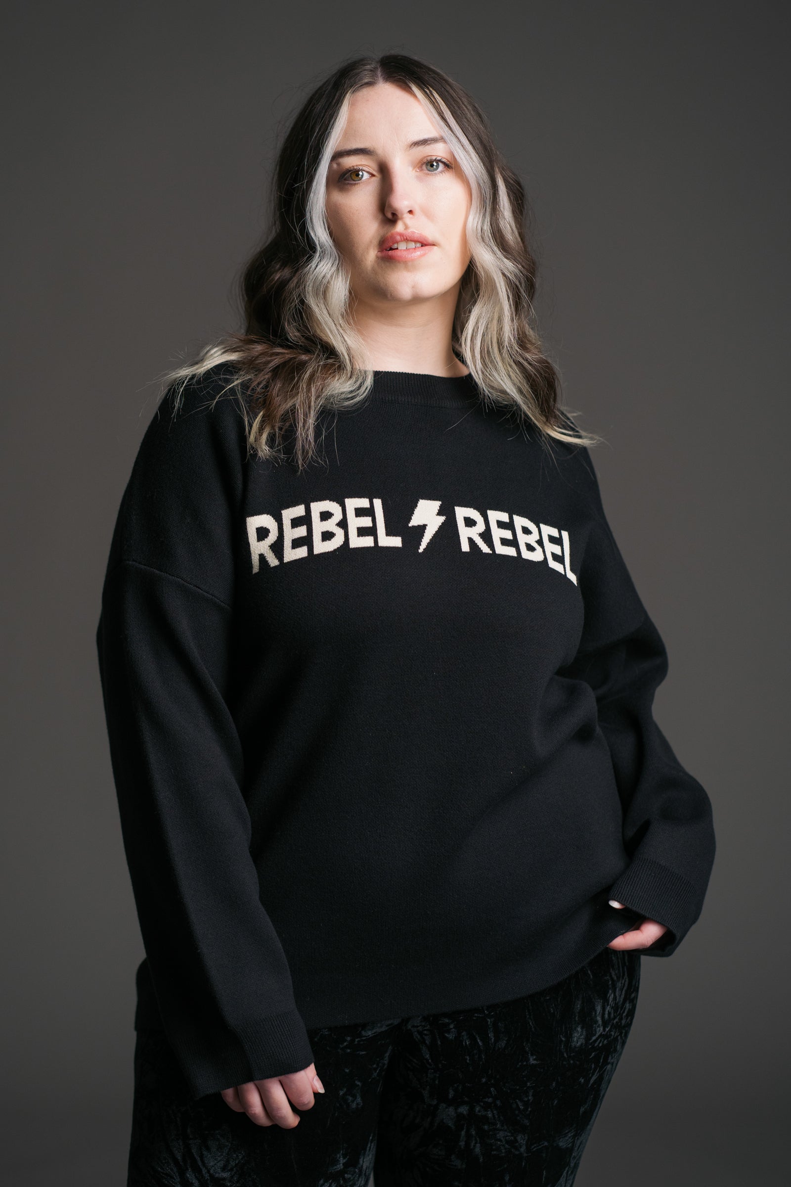 Taylor wearing a the Rebel Rebel Black Graphic Jumper with 'REBEL REBEL' ivory text and lightning bolt graphic on a gray background