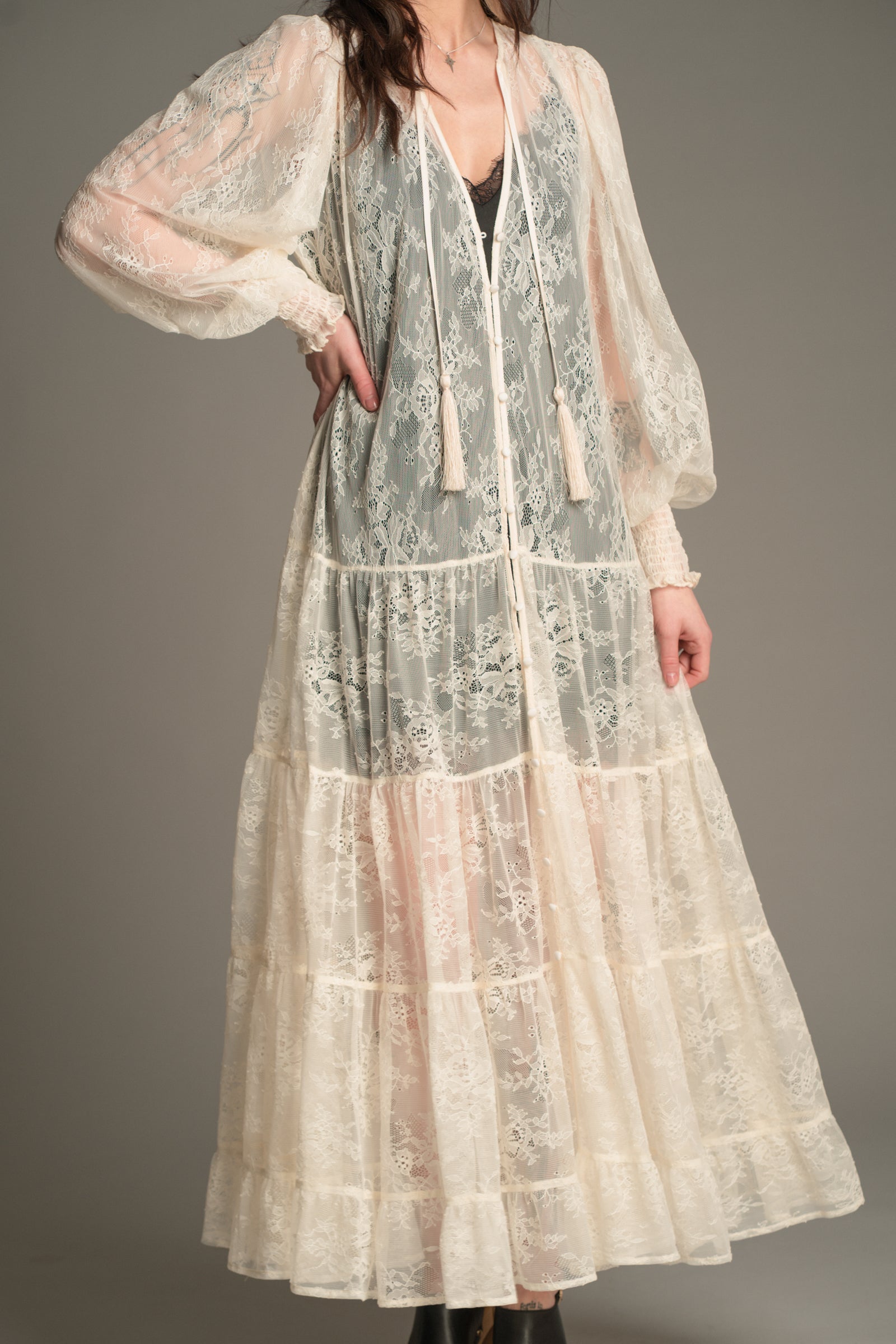 Front view of the Little Lies Rhiannon Ivory Maxi Dress featuring sheer ivory lace fabric, long voluminous sleeves, tassel ties, and a tiered maxi‑length skirt worn over the Nightbird Satin Slip Dress as an underlayer.