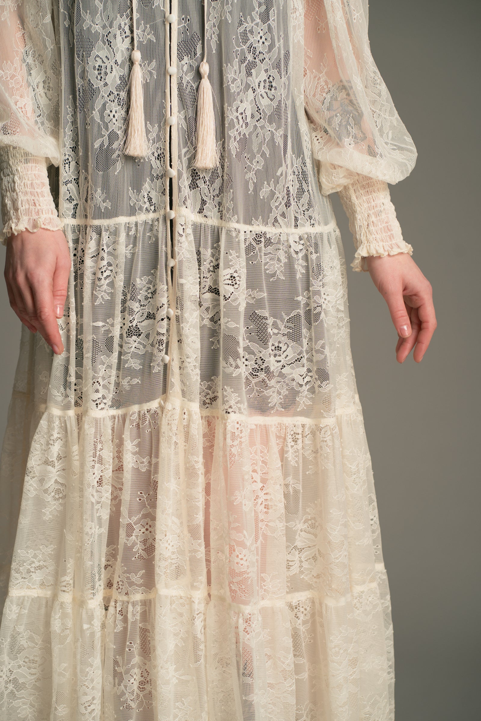 Close‑up detail of the Little Lies Rhiannon Ivory Maxi Dress showing sheer ivory lace fabric, tiered skirt panels, fabric‑covered buttons, and tassel ties.
