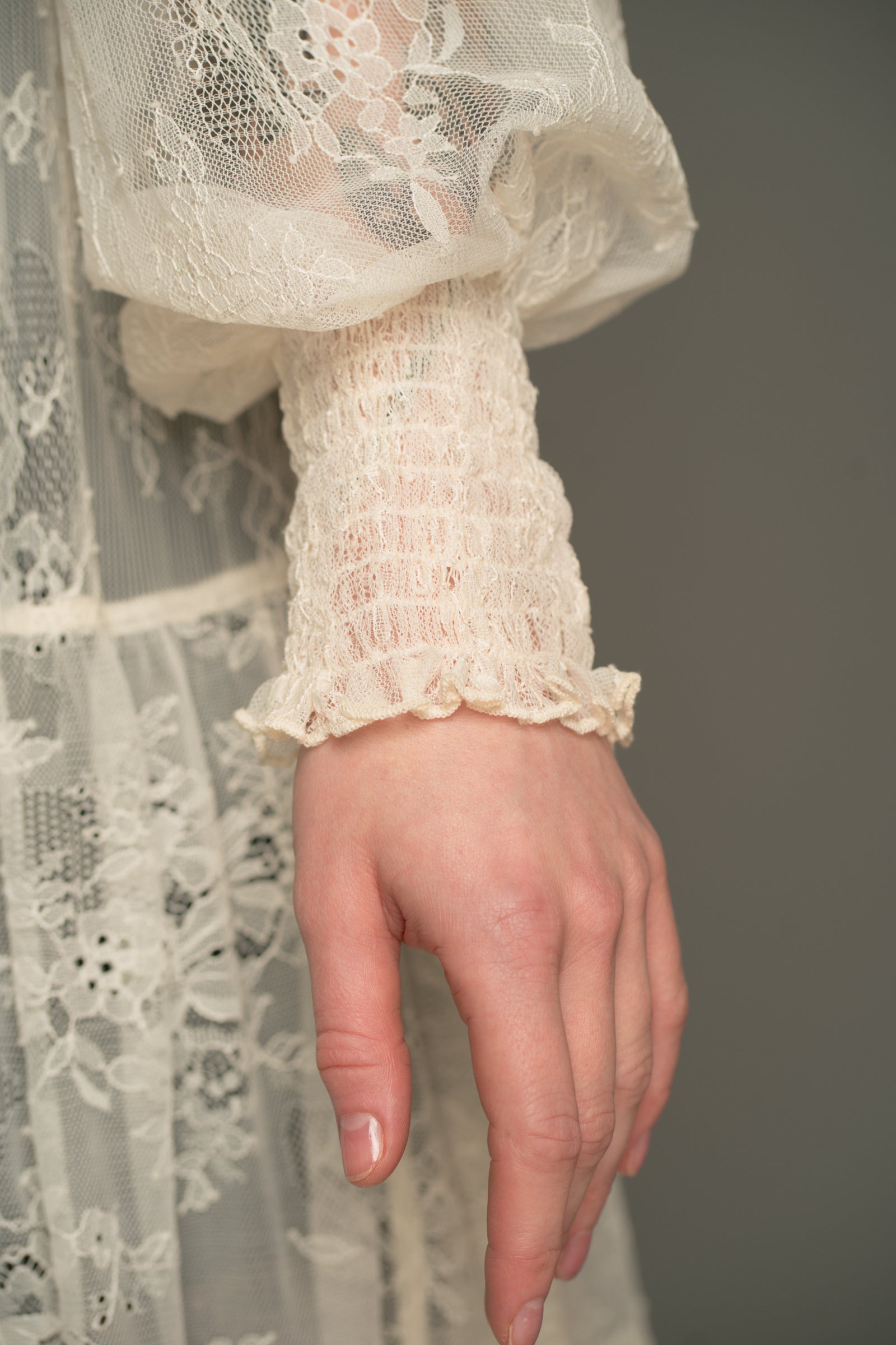 Close‑up detail of the Little Lies Rhiannon Ivory Maxi Dress showing the sheer ivory lace sleeve with a shirred cuff and the intricate lace panels of the skirt.